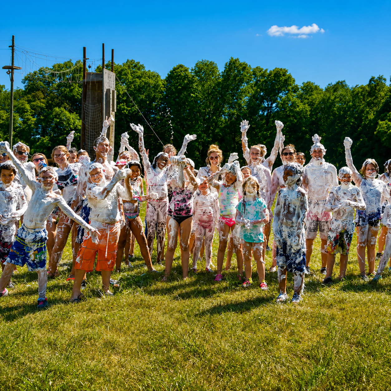A group of children and teenagers outdoors on a sunny day, covered in white foam or paint, celebrating and smiling with raised hands, wearing summer clothing, with a green tree background and a blue sky.