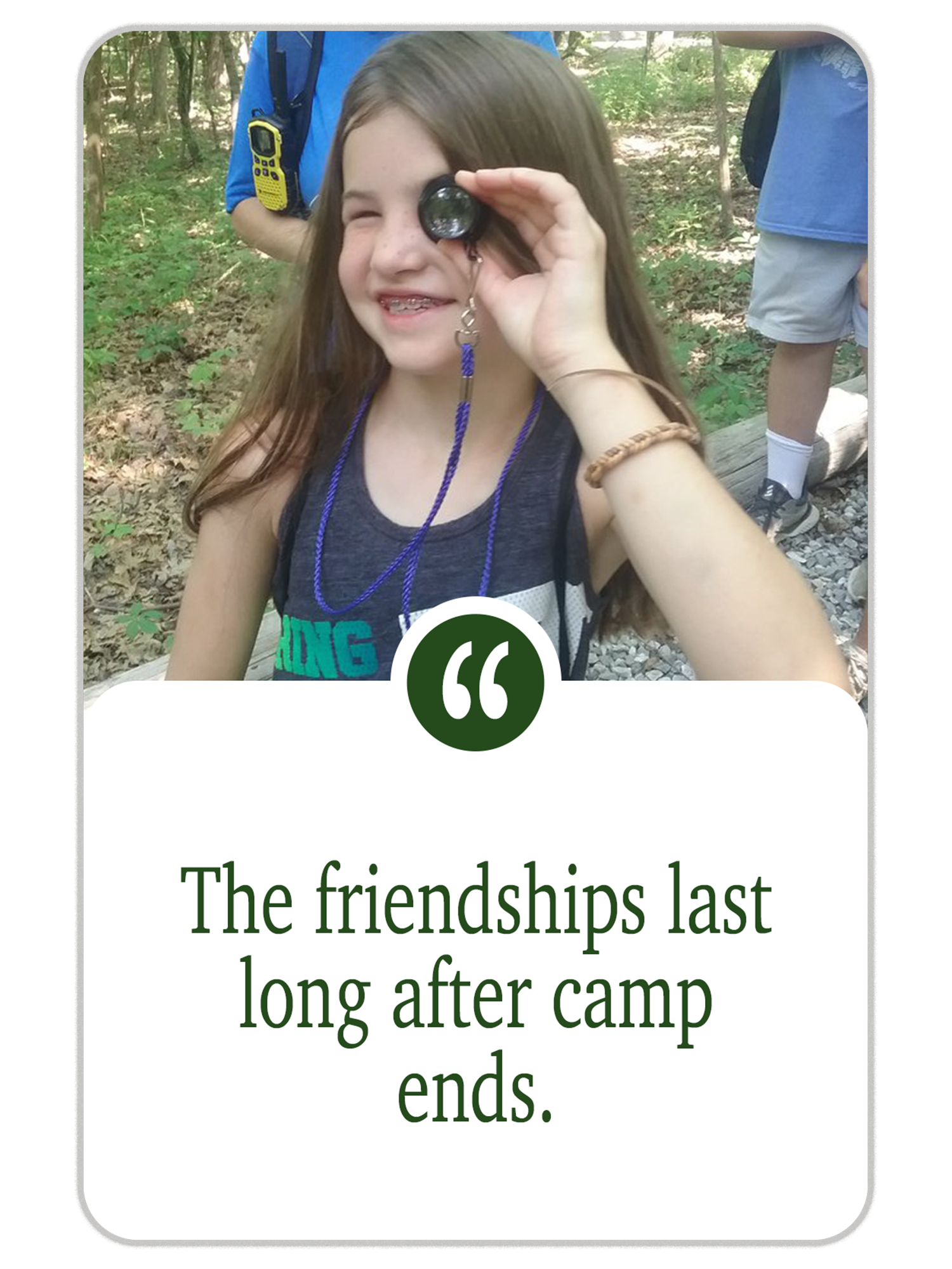 Young girl with a hand-held magnifying glass in her eye, smiling, in a wooded area with other children around.