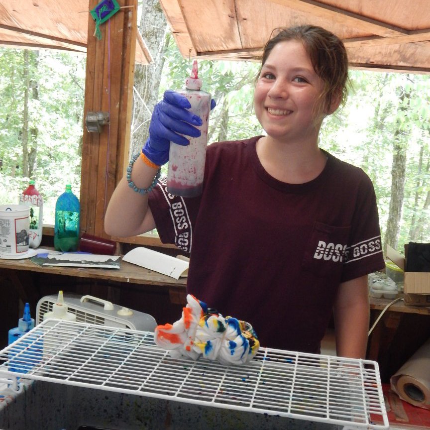 A young girl with short brown hair and a maroon t-shirt, smiling and holding a squeeze bottle of red and black tie-dye in an outdoor setup, with colorful tie-dye cloth on a table and bottles in the background.