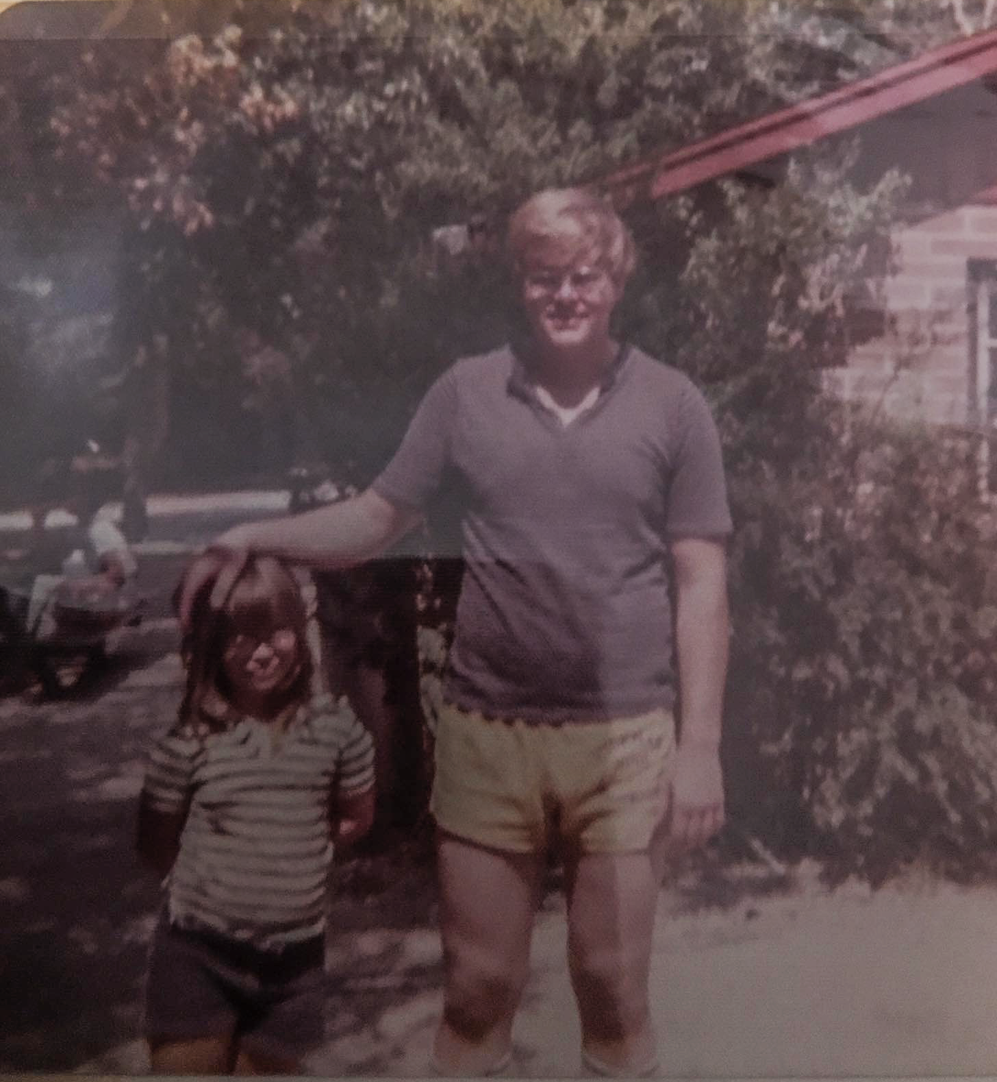A person with short blonde hair, wearing a gray T-shirt and yellow shorts, and a young girl with curly hair wearing a striped shirt standing outdoors in front of a tree and a house, with a person on a motorcycle in the background.