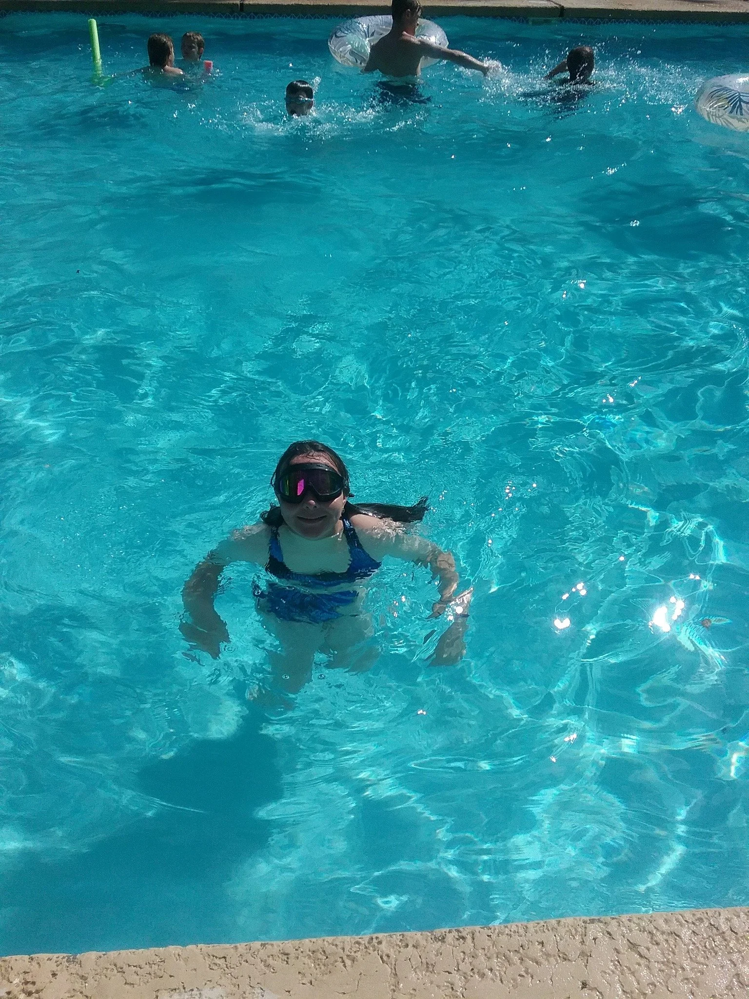 People swimming and playing in a bright blue swimming pool, with a person in goggles in the foreground.