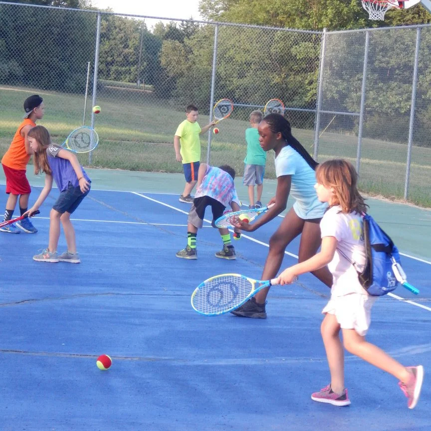 Children playing tennis on a blue outdoor tennis court with a chain-link fence and trees in the background.