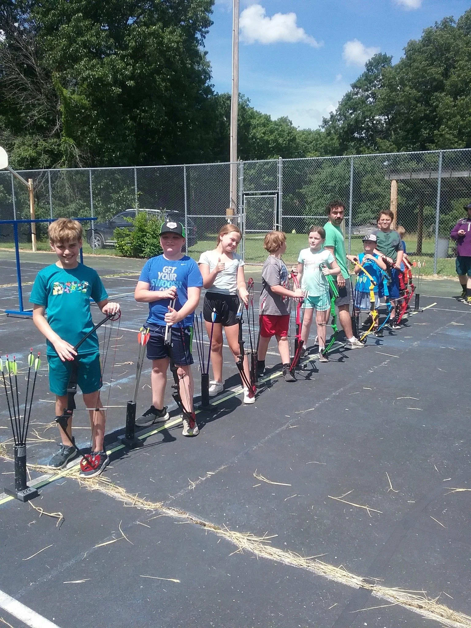 A group of children and two adults standing in a line on a basketball court, holding bows and arrows, preparing to shoot. There is a chain-link fence in the background and green trees surrounding the area.