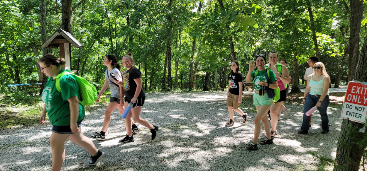 Group of young women and girls hiking on a forest trail, some smiling and waving, surrounded by green trees, with a sign indicating 'Exit Only' and 'Do Not Enter' near trees.