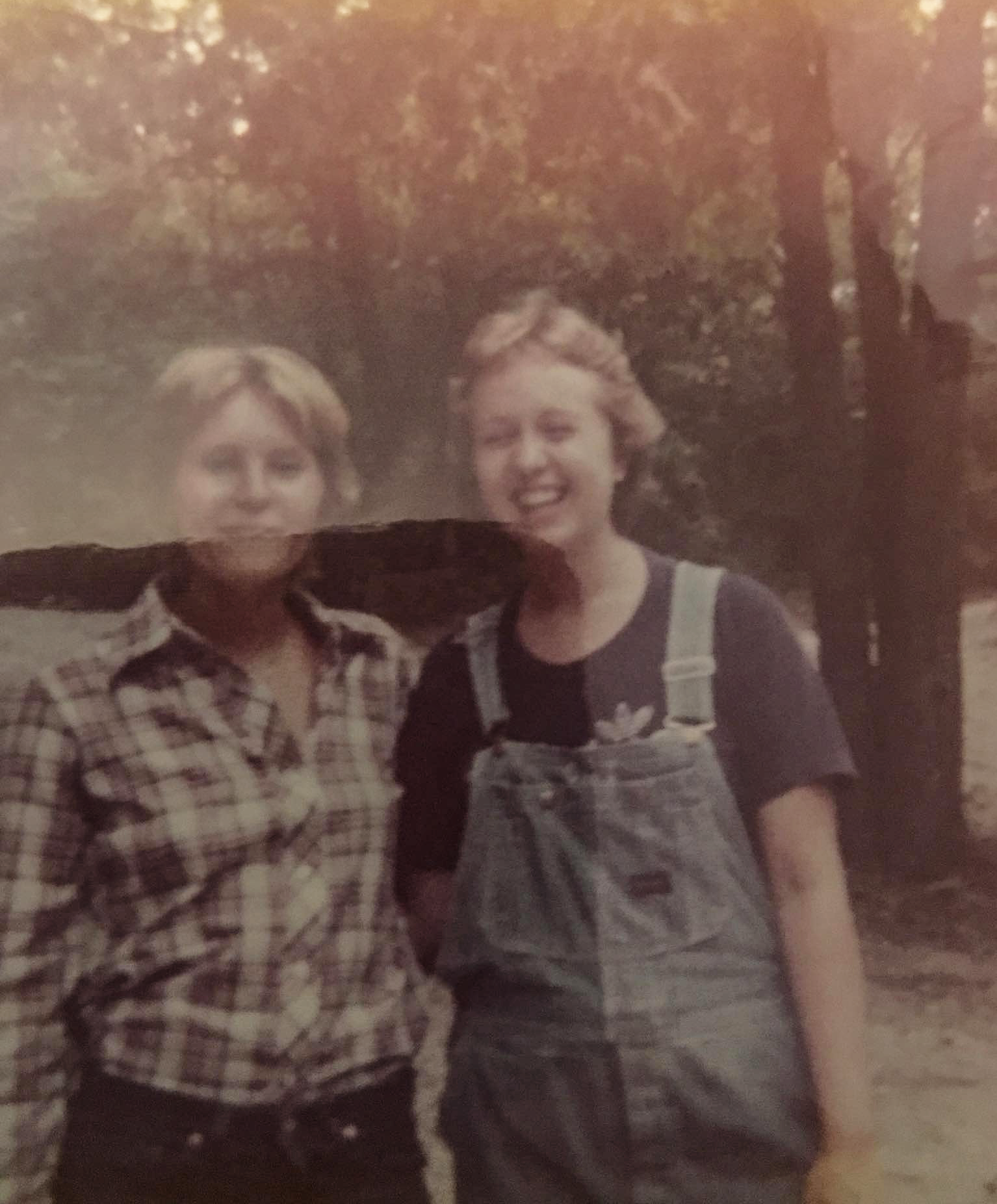 Two women standing outdoors in front of trees, smiling at the camera. The woman on the left has short blonde hair and is wearing a checkered shirt. The woman on the right has curly blonde hair and is wearing a dark T-shirt with overalls.