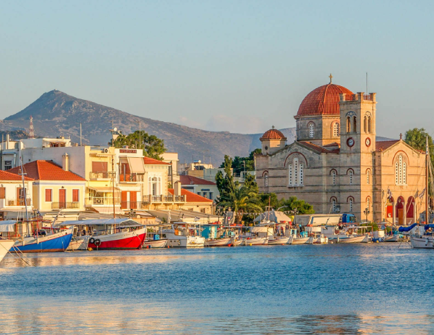 A harbor with boats docked along the water, a church with a large dome and cross, colorful buildings, and mountains in the background.