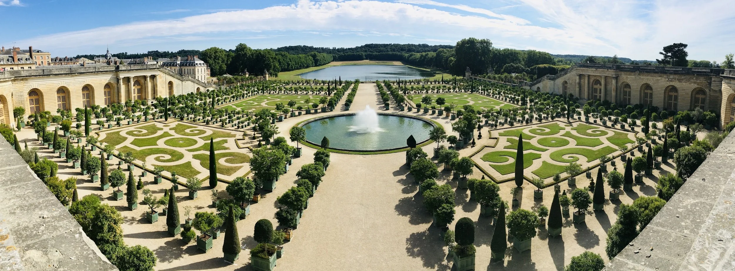 Panoramic view of the formal French garden with geometric patterns, a fountain in the center, symmetrical trees, and a large pond at the background under a blue sky with some clouds.