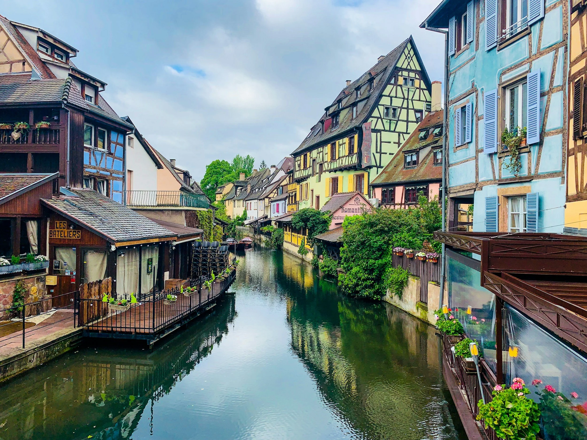 Colorful European-style buildings with shutters line a small canal with still water, greenery, and a partly cloudy sky.