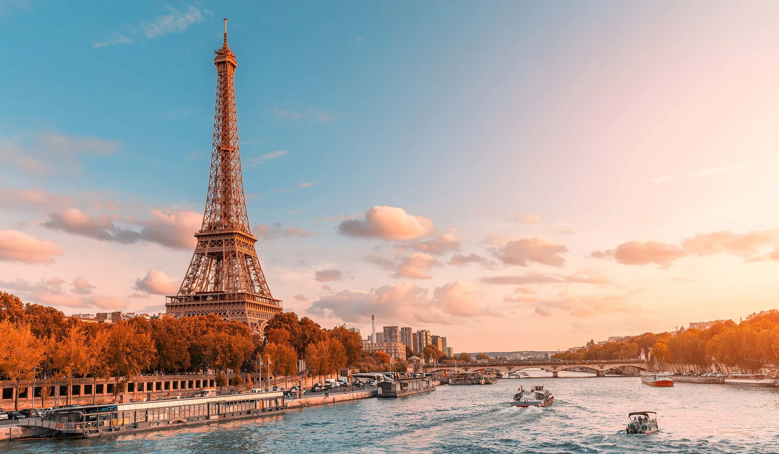 Eiffel Tower in Paris, France, viewed from across the river during sunset, with boats on the water and buildings in the background.