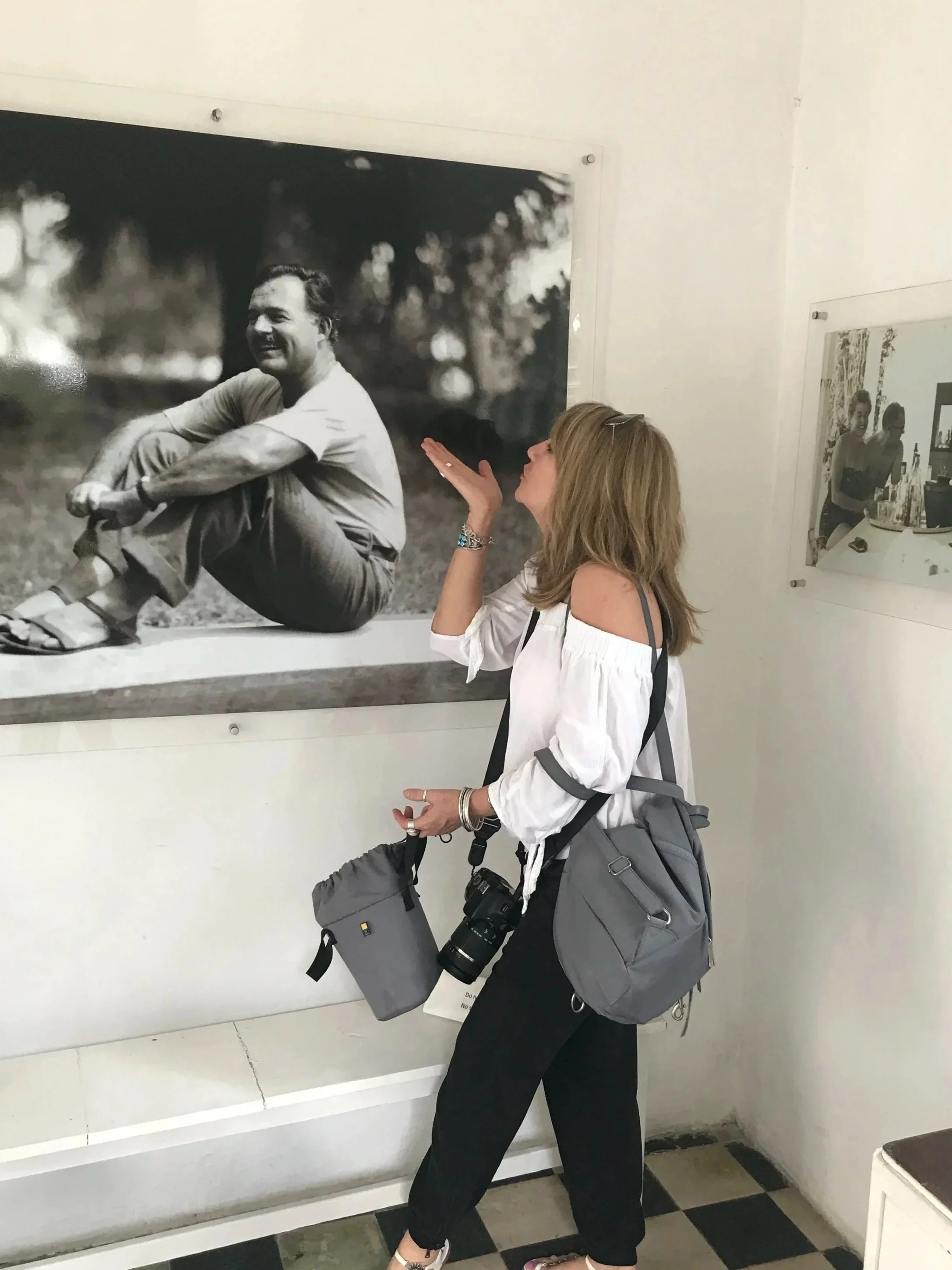 A woman with long blonde hair wearing a white off-the-shoulder top and black pants is standing in front of a black-and-white photograph, appearing to blow a kiss or make a gesture towards it. She has a large gray backpack with a camera hanging from it, and is wearing sandals and jewelry.