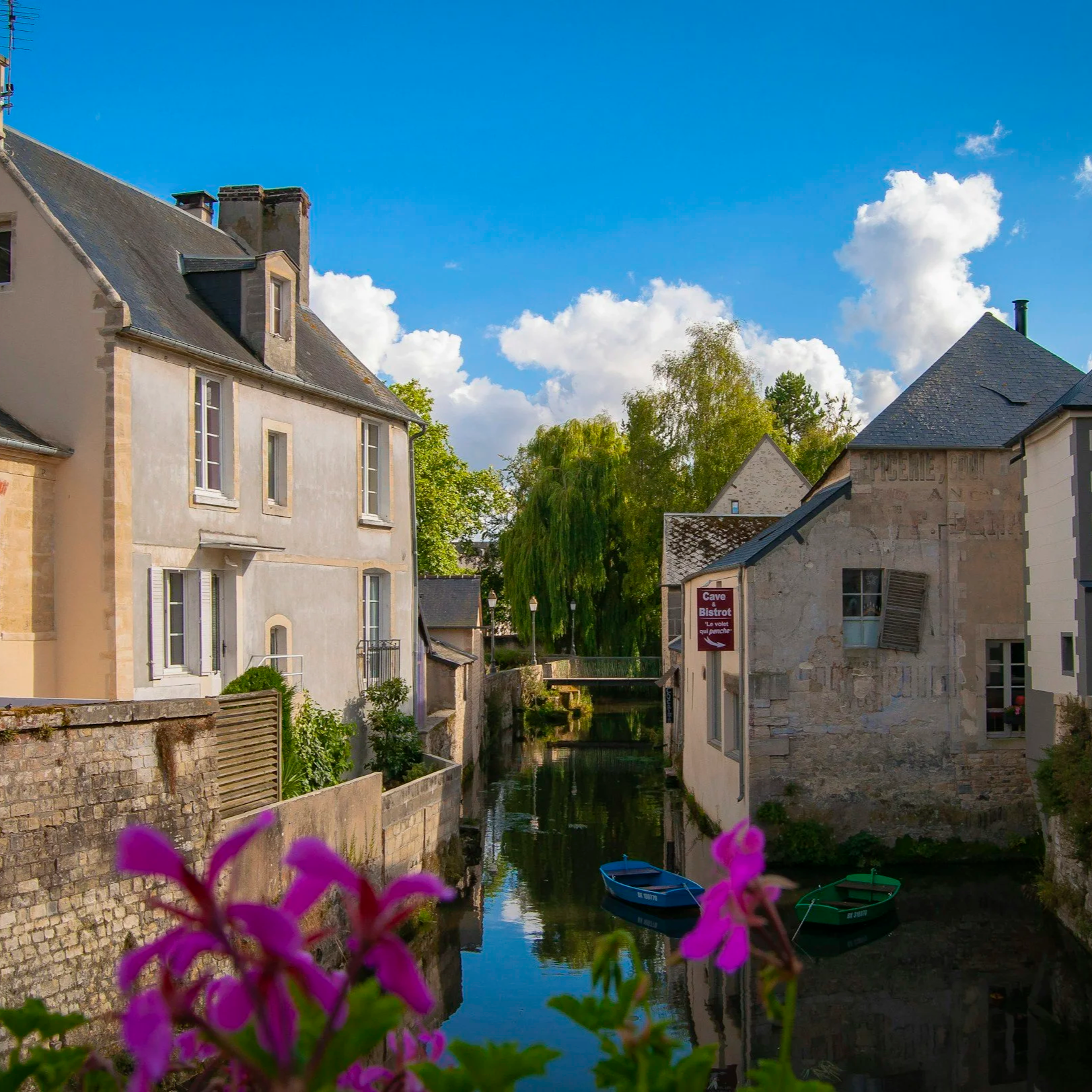 View of a small canal lined with stone and brick buildings on a sunny day with a bright blue sky and white clouds. Two small boats are floating in the canal, and purple flowers are in the foreground.