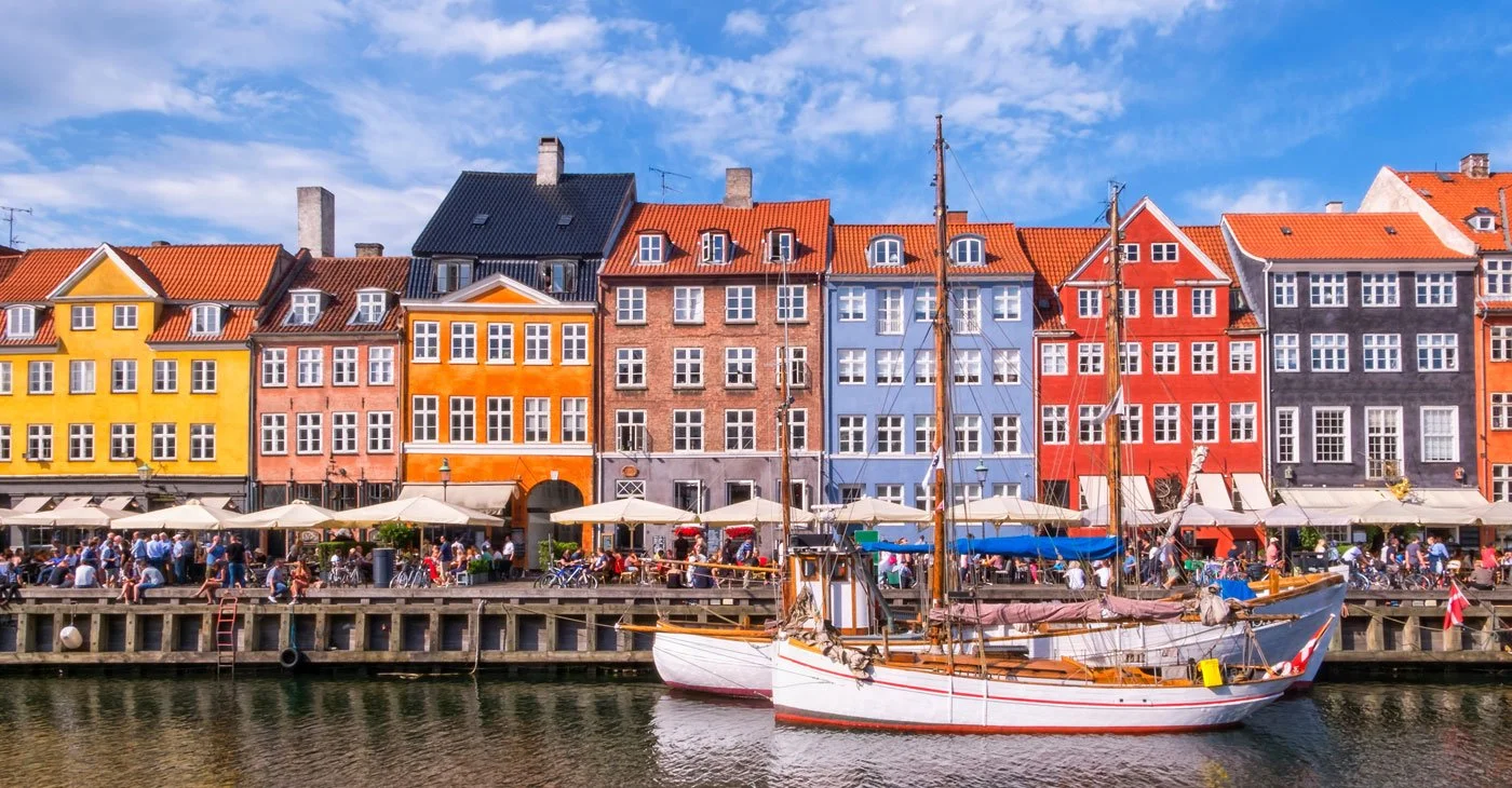Colorful buildings along a waterfront with boats docked in the water and people dining at outdoor cafes.
