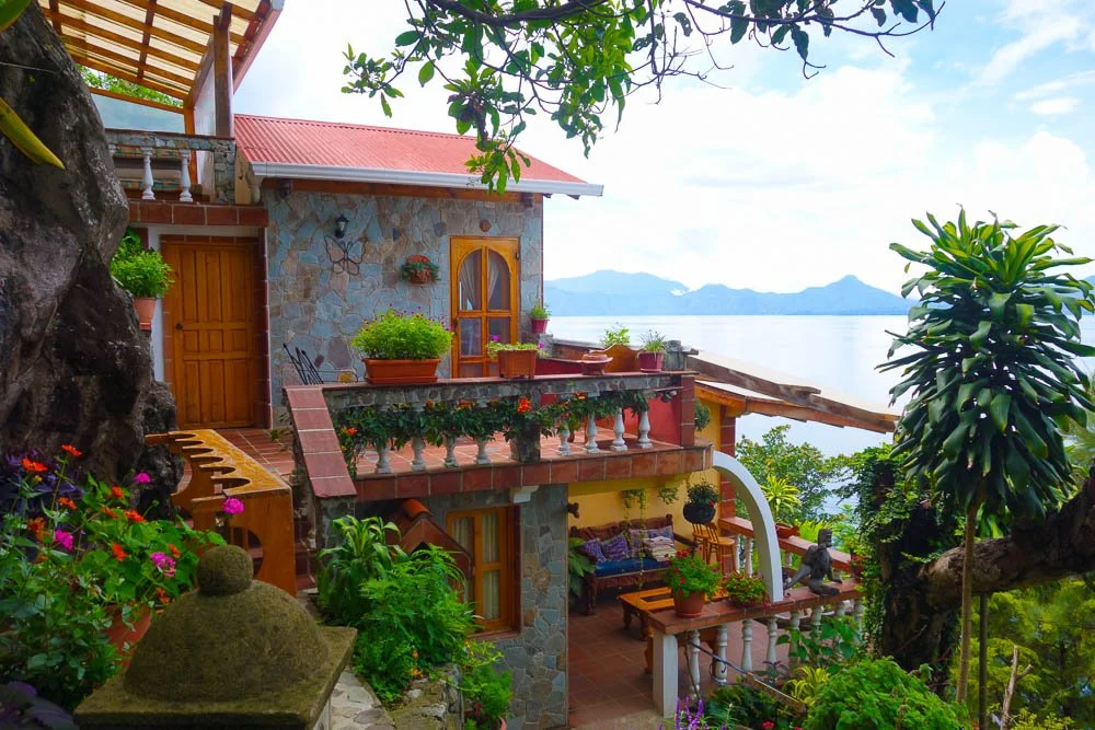 A multi-level house with stone walls and wooden doors, surrounded by lush greenery and colorful flowers, overlooking water and mountains in the distance.