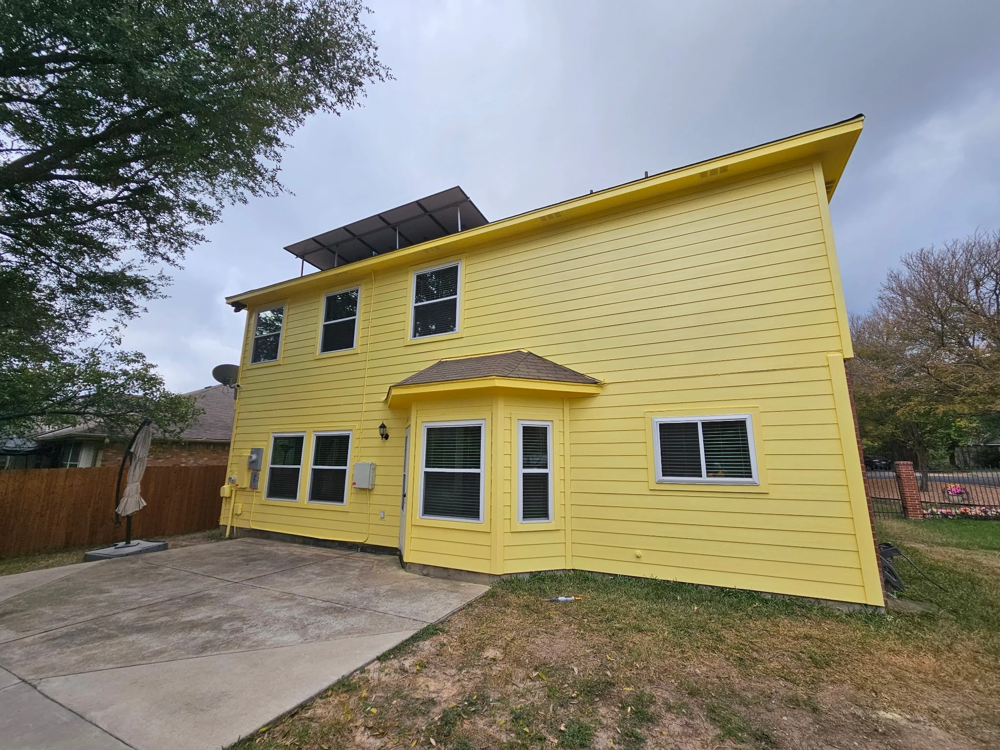Yellow two-story house with multiple windows, a small porch, and solar panels on the roof, surrounded by trees and a fenced backyard.