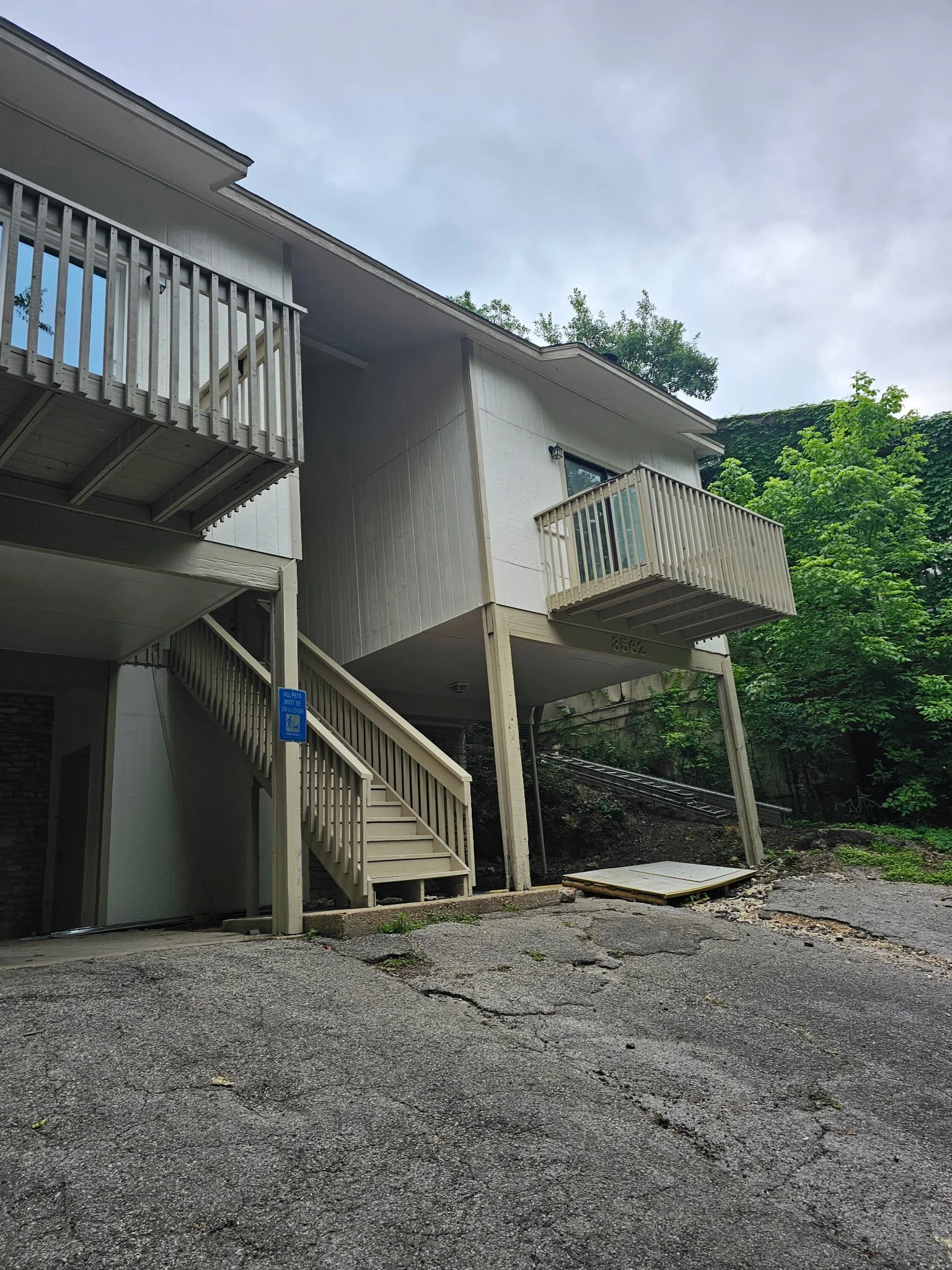 Apartment building with beige exterior and balconies, surrounded by green trees, with an uneven asphalt area in the foreground. Siding and trim replacement. exterior paint job.