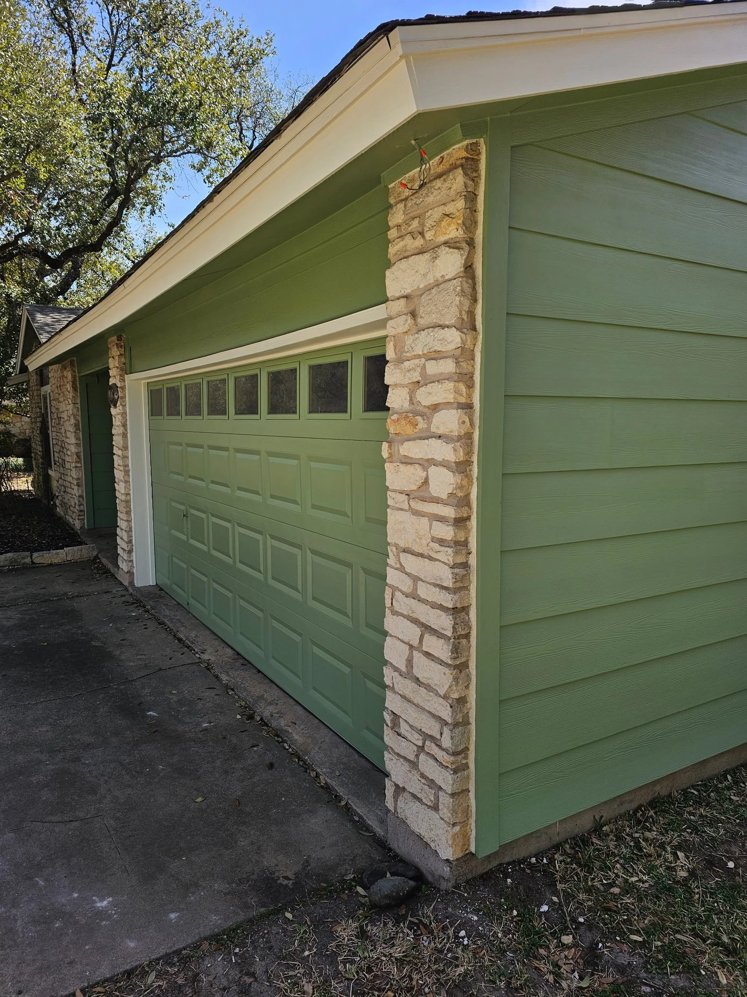 The image shows the corner of a house with green siding, a white gutter, and a stone column. There are small windows on the garage door, and the background includes trees and a blue sky.