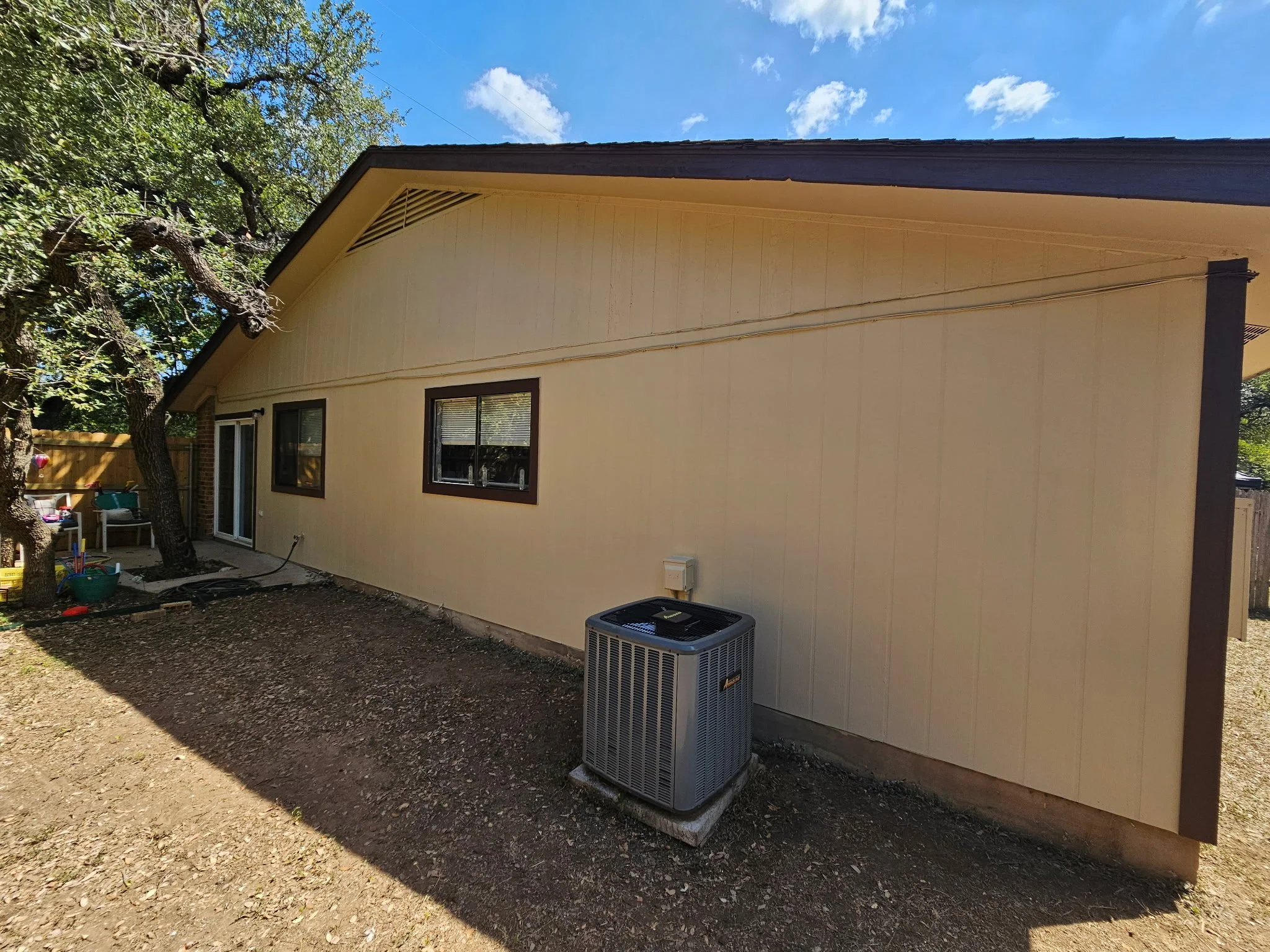 The back of a beige house with two black-framed windows, a sliding glass door, and a tree in the yard. An air conditioning unit is installed at the base of the house. The sky is blue with scattered clouds.