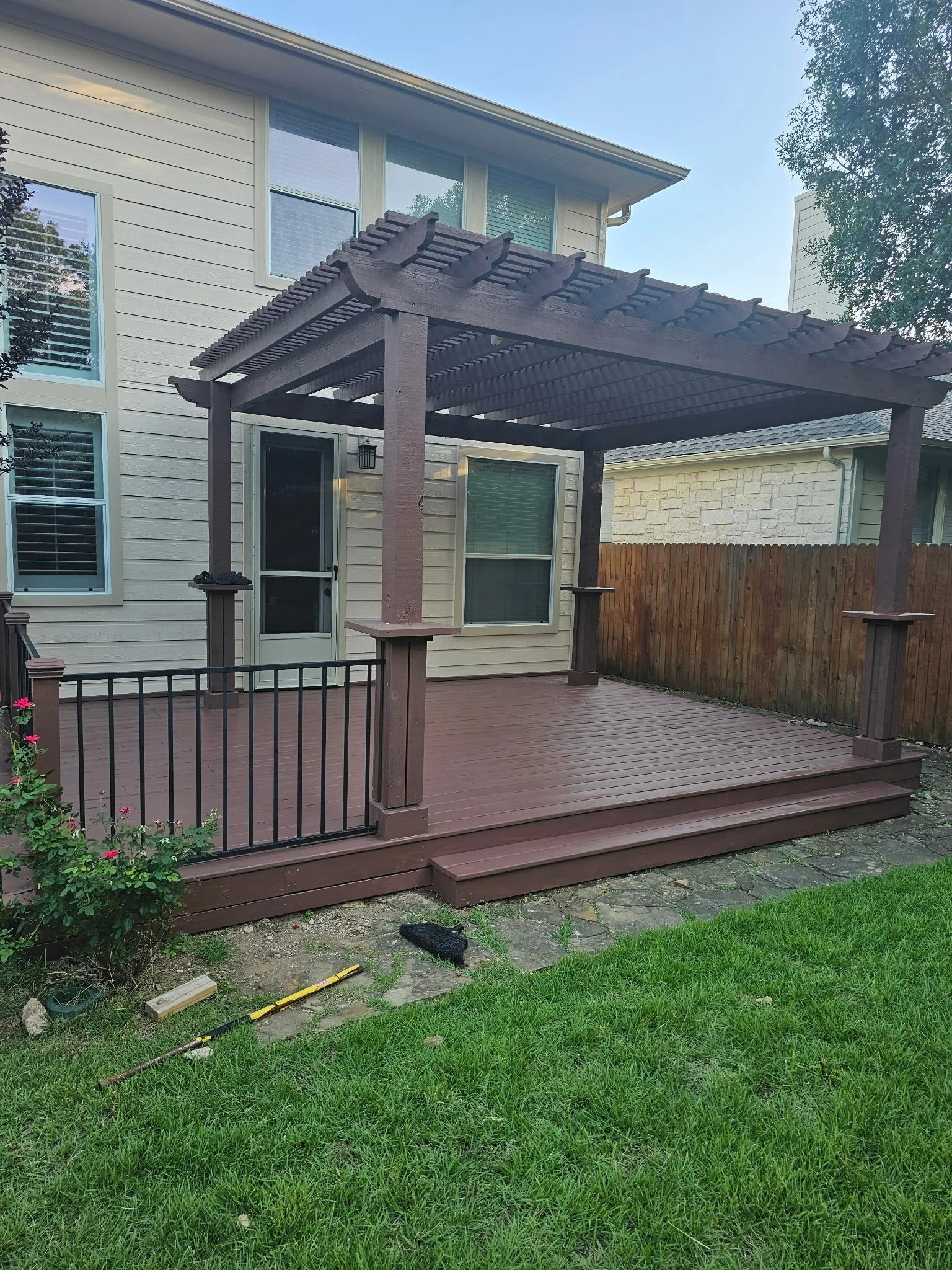 A backyard deck with a pergola attached to a beige house with white trimmed windows and door. The deck has steps leading down to grass, a black metal railing on one side, and construction tools and materials scattered on the ground.