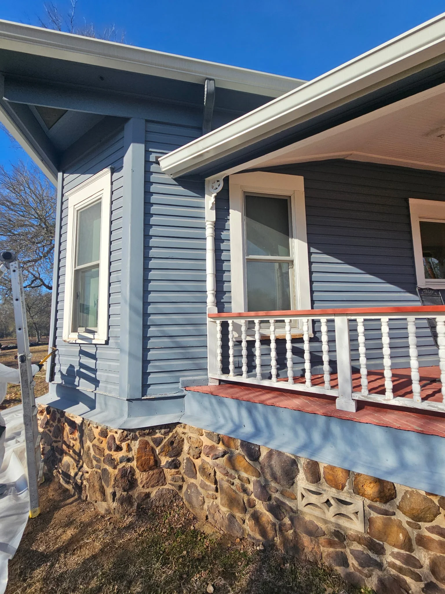 View of the corner of a house with blue siding, white trim, and a stone foundation. There is a stone wall base, a small porch with a white railing, and a window with white trim. The roof has guttering and the sky is clear and blue.