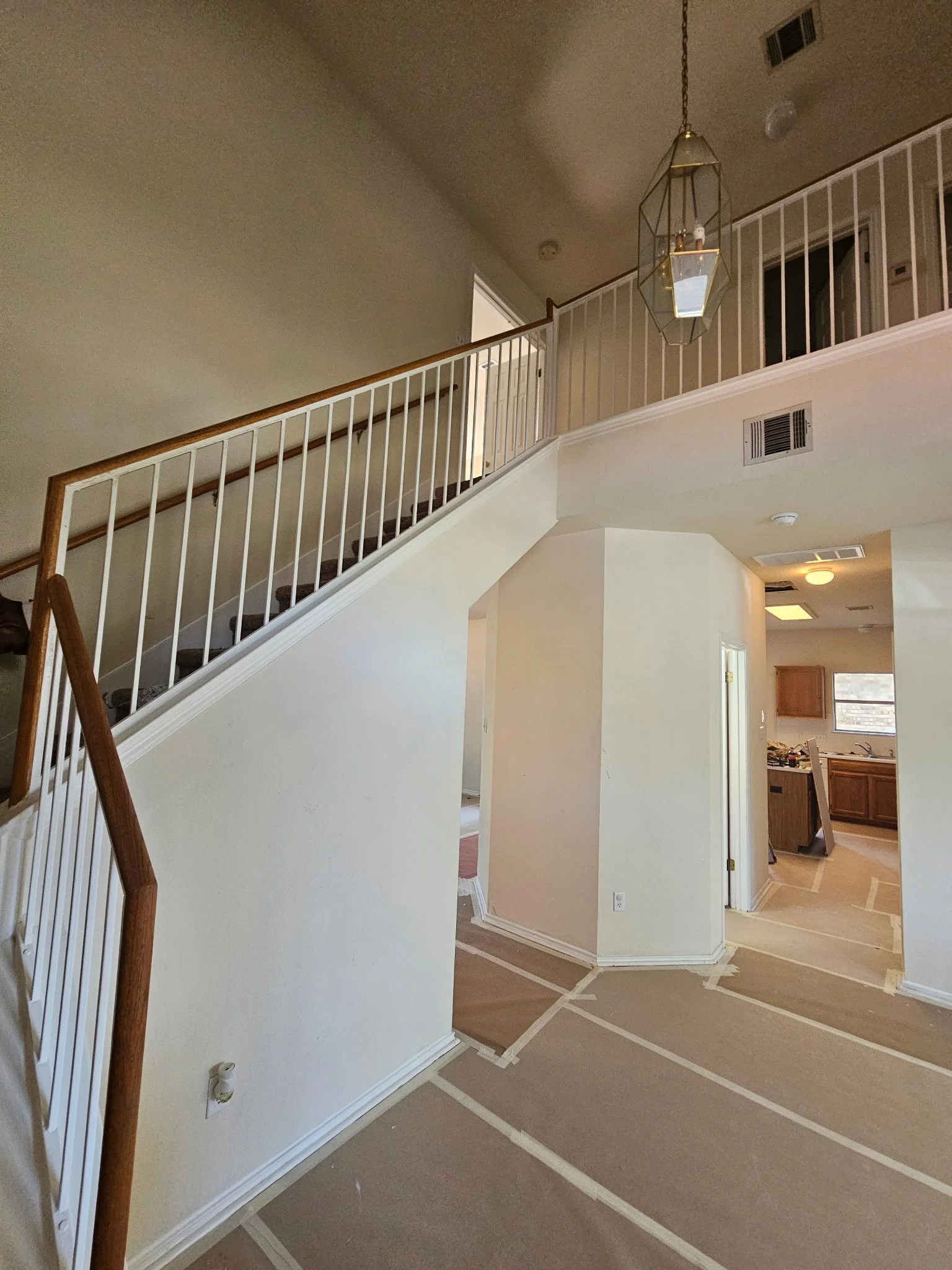 The image shows a view of the interior of a house undergoing renovation, with taped-off areas on the floor and walls, to protect surfaces during painting or flooring work. There is a staircase with a white railing and wood handrail, a hanging geometric light fixture, and part of a kitchen with wooden cabinets and a window.