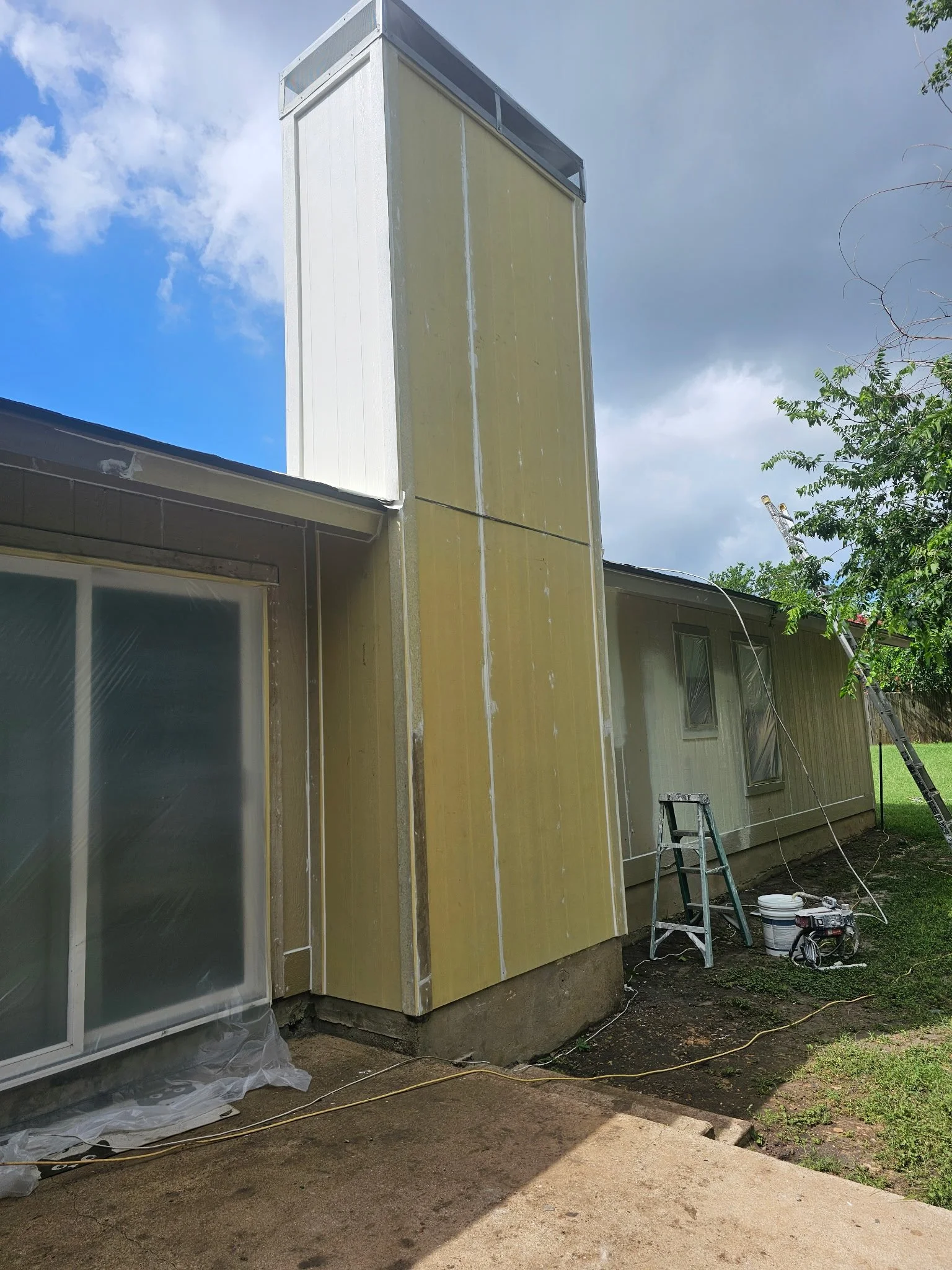 Side of a house under construction with yellow insulation siding, windows, ladders, and construction tools, and trees in the background.