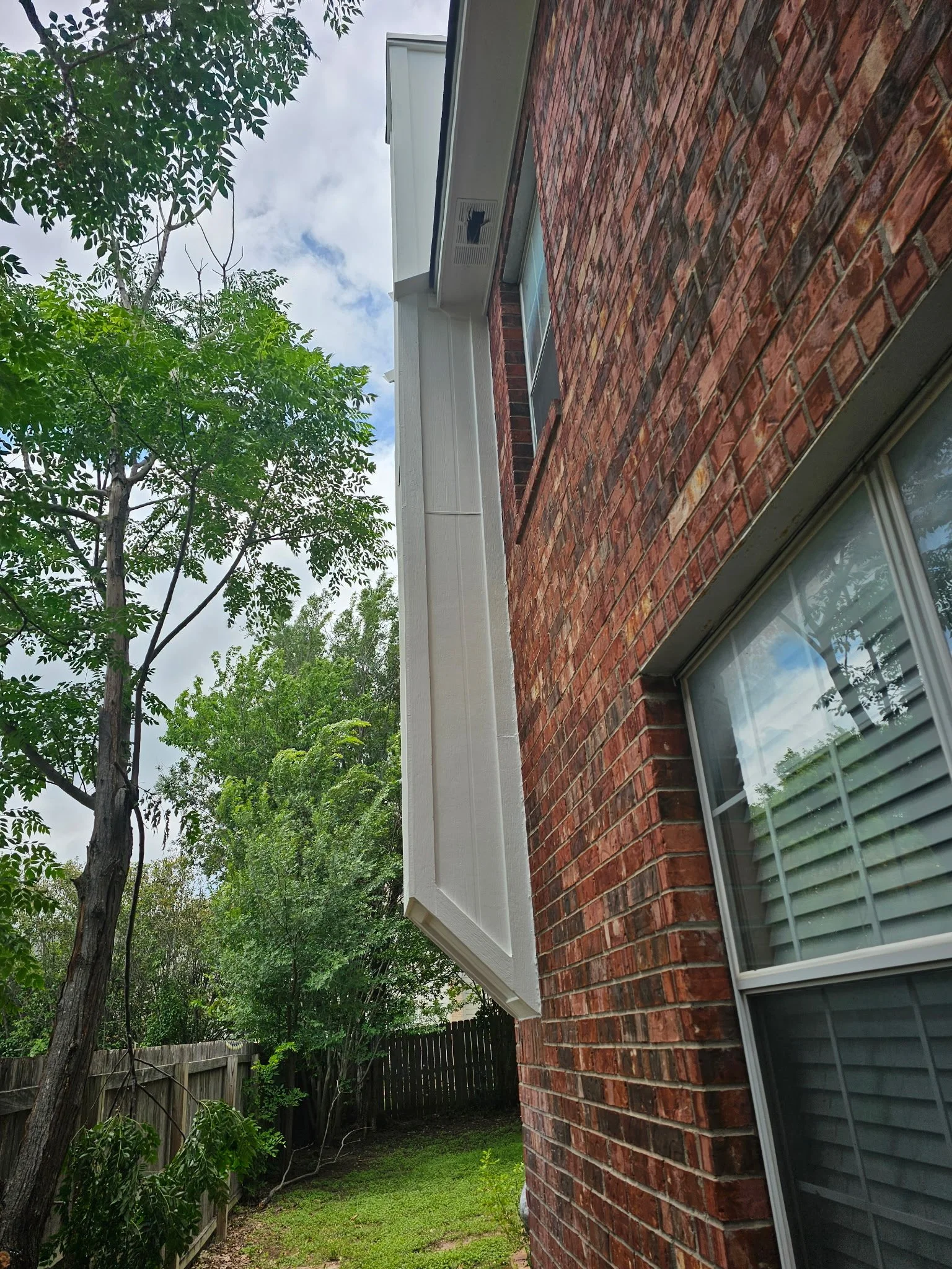 View of the side of a brick house with a large window and white trim, surrounded by green trees and a backyard with grass and a wooden fence.