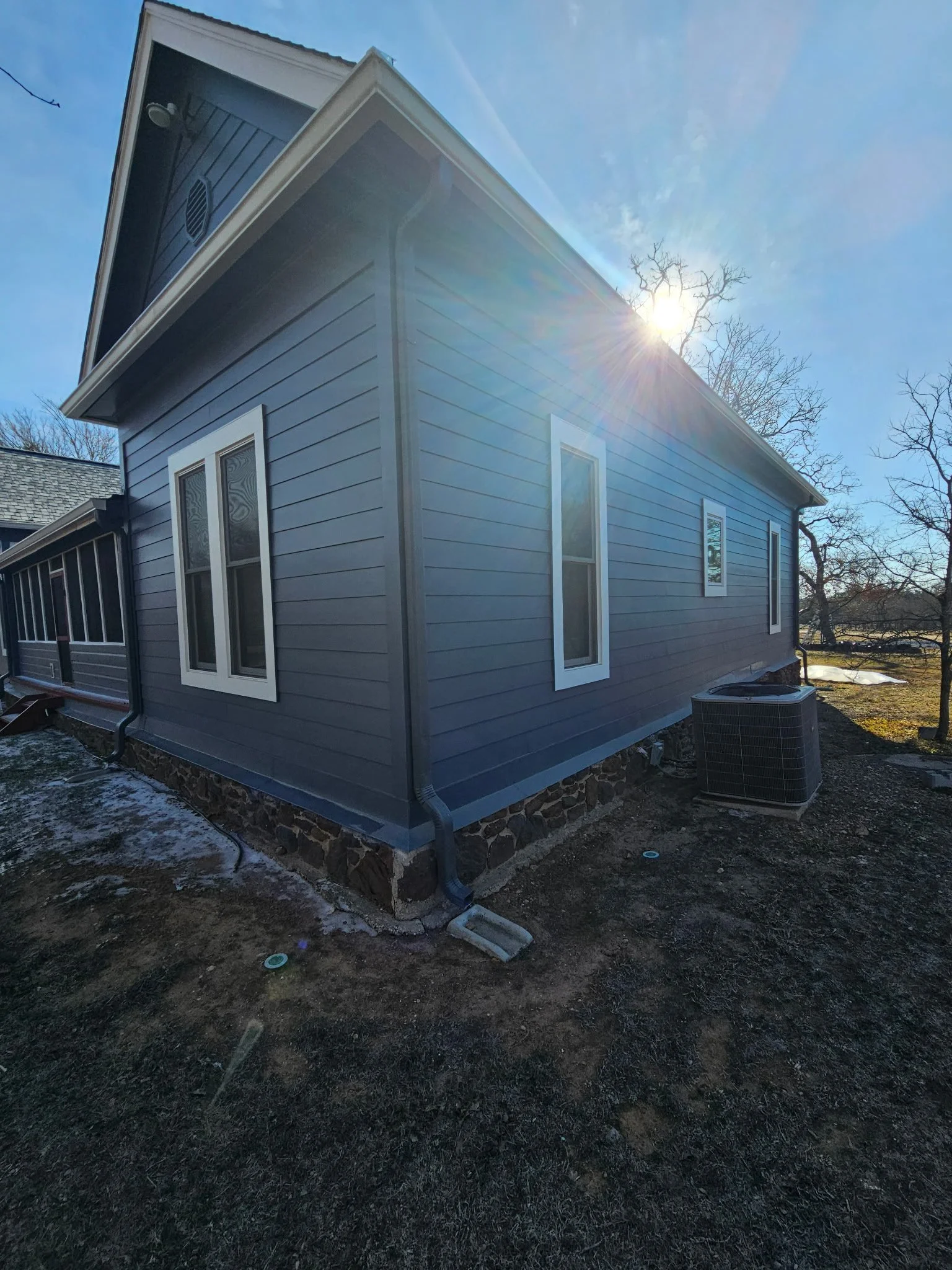 The image shows the side view of a house with gray siding and white trim, with three windows along the side. There is a vent and a small light on the house's upper part. An HVAC unit is present at the back, and the ground appears to be dirt with some patches of snow. The sky is clear with the sun shining brightly behind a tree.