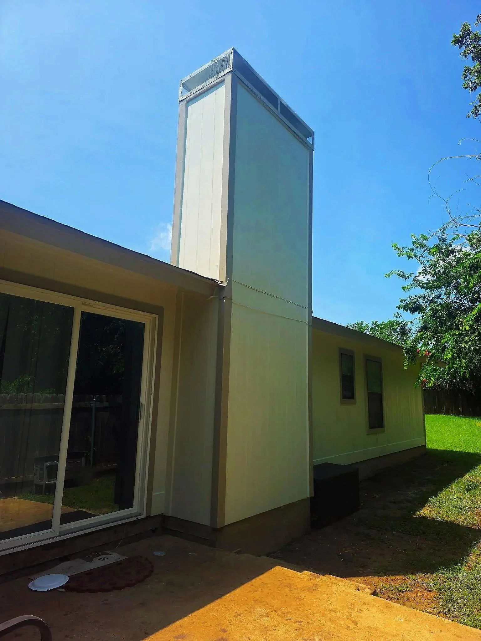The image shows the corner of a house with a large, white, rectangular extension or addition featuring windows. The sky is clear and blue, with a few trees visible on the right side. There are some small objects and paving materials on the ground near the house, indicating ongoing exterior work or landscaping.