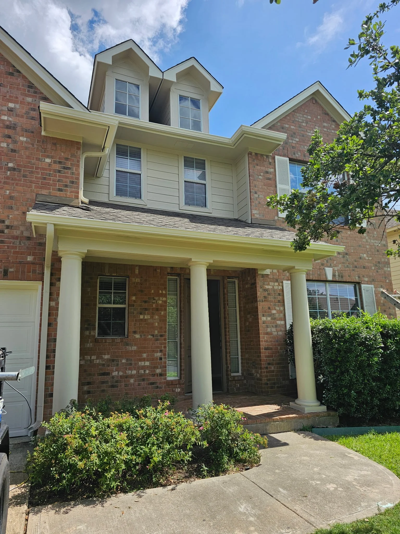 Front yard of a brick house with a covered porch supported by white columns, multiple windows, and green bushes and plants along the sidewalk. A tree is visible on the right side of the image, and part of a driveway is seen on the left.