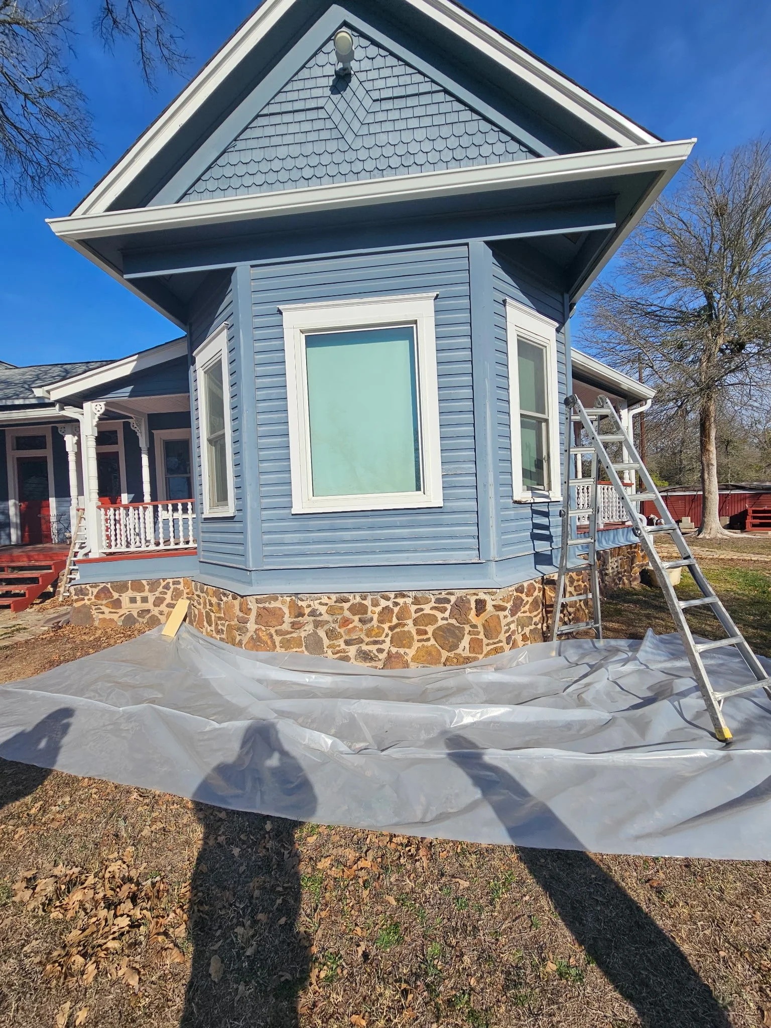 The image shows the exterior of a blue house with white trim and a stone foundation. A ladder leans against the house, and a white tarp is spread on the ground. The sky is clear and blue, and there are leafless trees in the background.