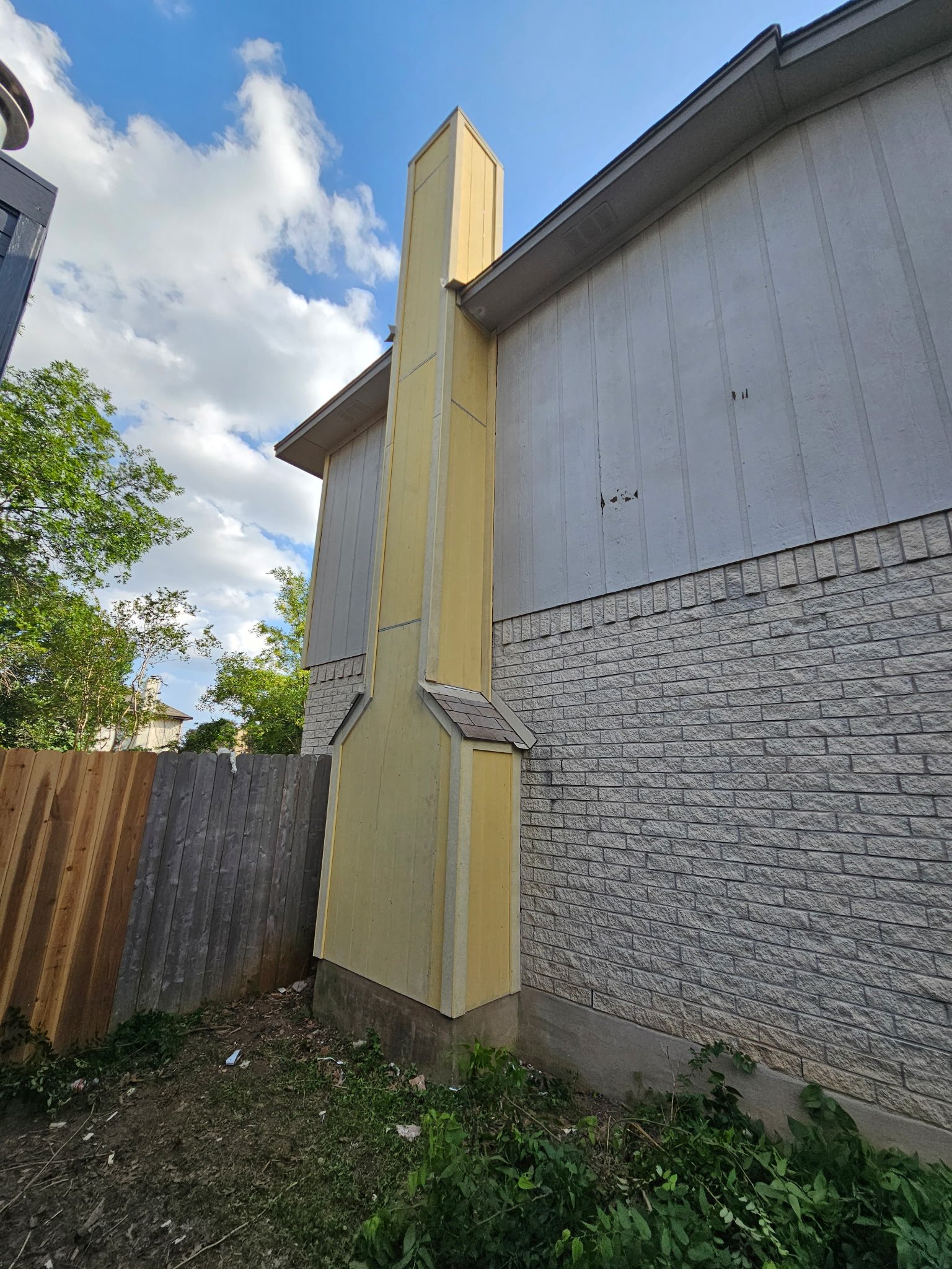 A house with a brick lower wall, white upper wall, and beige vertical section near the corner, with a sloped roof. Replacing siding with James Hardie fiber cement panA wooden fence runs along the left side, and there are bushes and grass at the base.