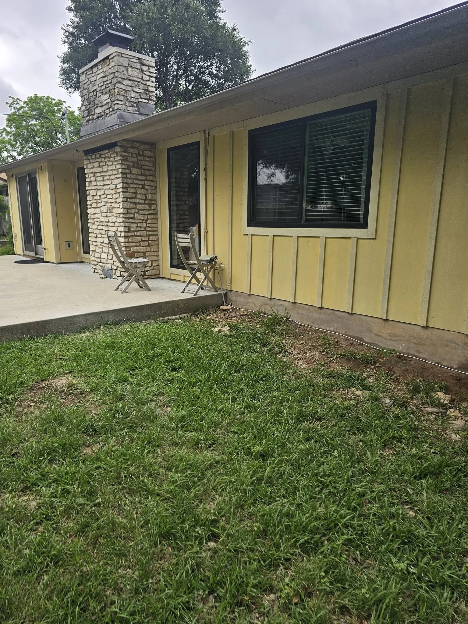 Side view of a single-story house with yellow exterior walls, large windows with blinds, a stone chimney, a small concrete porch with two chairs, green grass, and trees in the background under a cloudy sky.