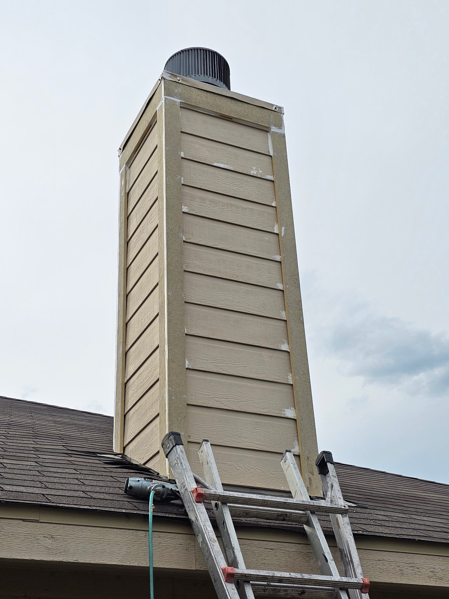 A ladder leaning against a house with scaffolding around a chimney, which is under construction or repair, on a roof with shingles, siding repairs and trim repairs with a cloudy sky in the background.