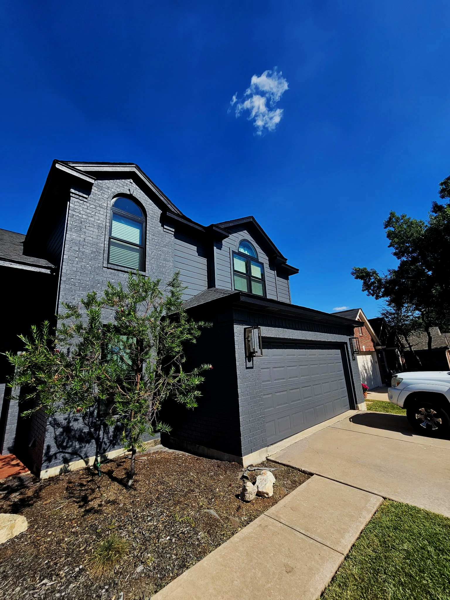 A modern house with a gray exterior, arched windows, and a garage, surrounded by trees and a sidewalk under a blue sky with a small cloud.