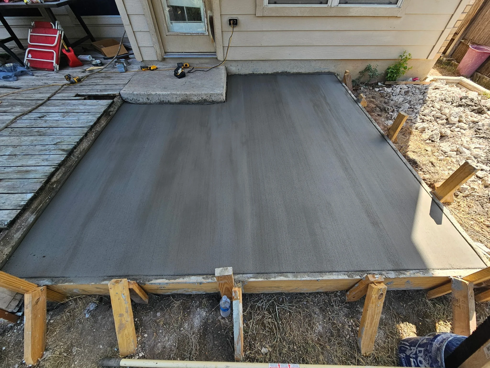 A freshly poured concrete slab forms the foundation for a new deck or porch attached to a house with beige siding. Wooden framing supports the concrete, and construction tools and materials are scattered nearby.