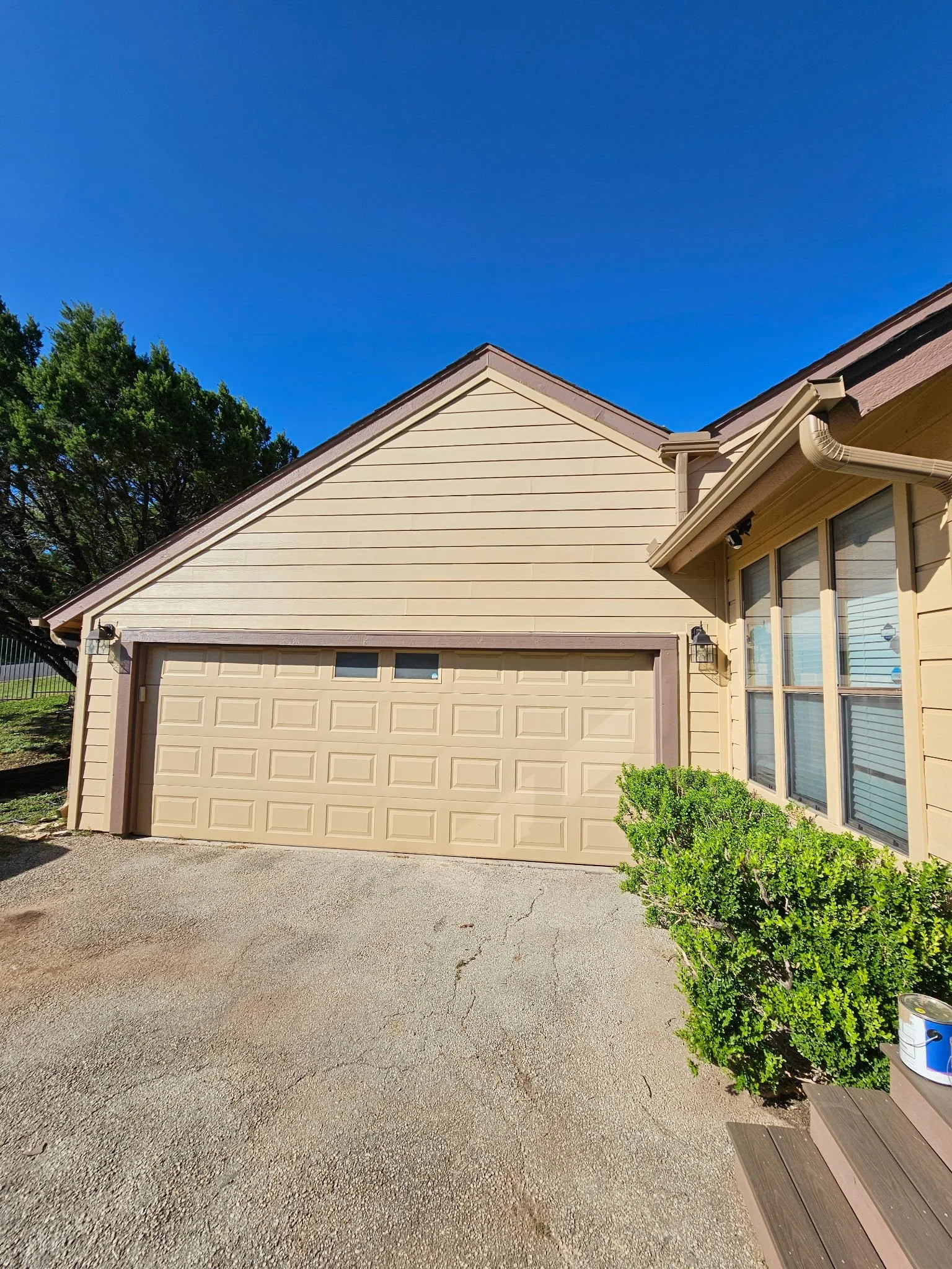 A house with a beige garage door, tan siding, and large windows. A green shrub and a wooden bench are in the front yard, with a bright blue sky above.