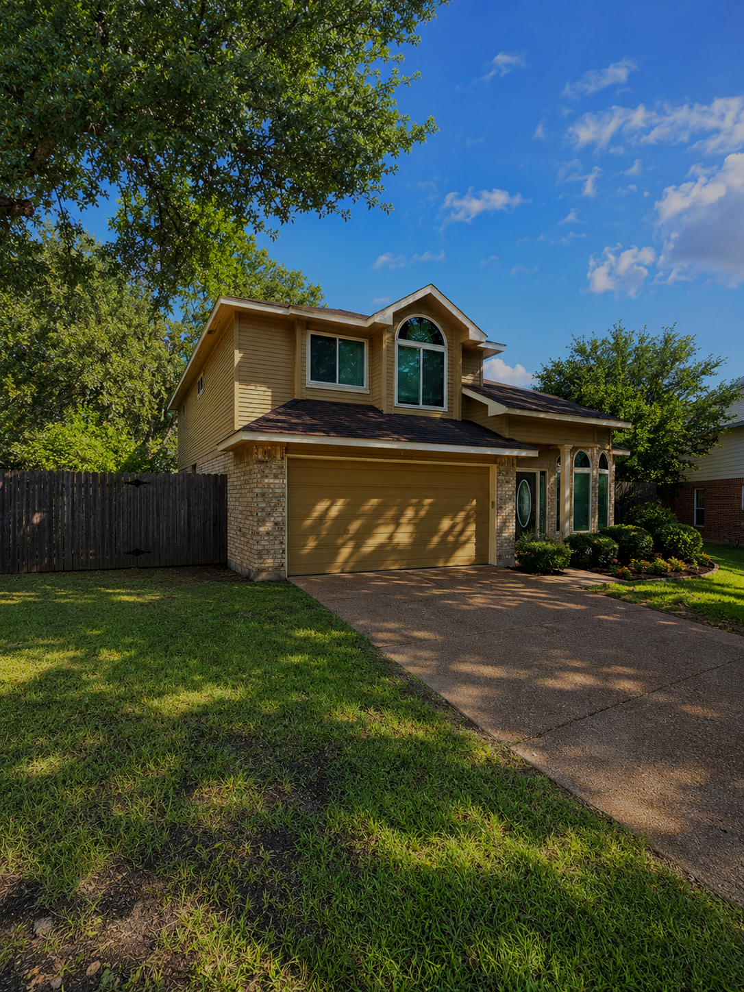 Two-story suburban house with yellow siding, brick accents, and a two-car garage, surrounded by green lawn and trees, under a blue sky with some clouds.
