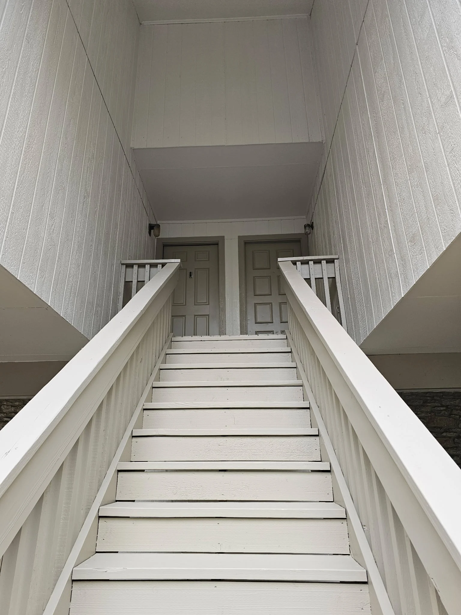 White wooden staircase leading to a landing with a gray door at the top. The staircase is flanked by white wooden railing, and the walls are paneled in white wood.