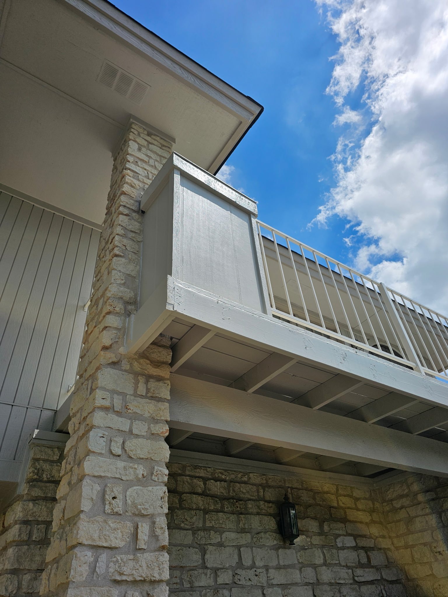 Exterior view of a building with stone and white siding, a balcony with white railings, and a blue sky with clouds.
