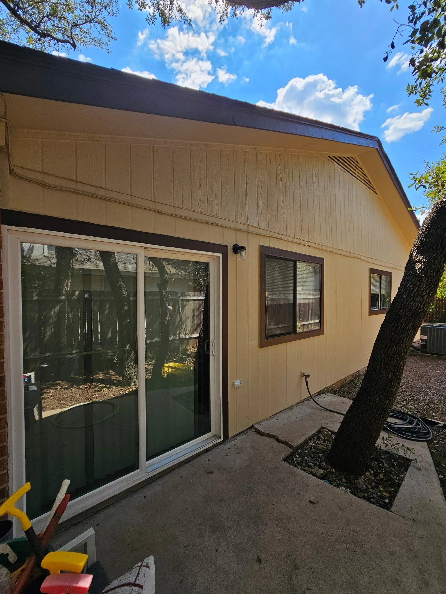 Backyard view of a house with beige walls, dark brown window frames, and a sliding glass door. There is a tree trunk on the right side with branches overhead. The sky is partly cloudy and blue.