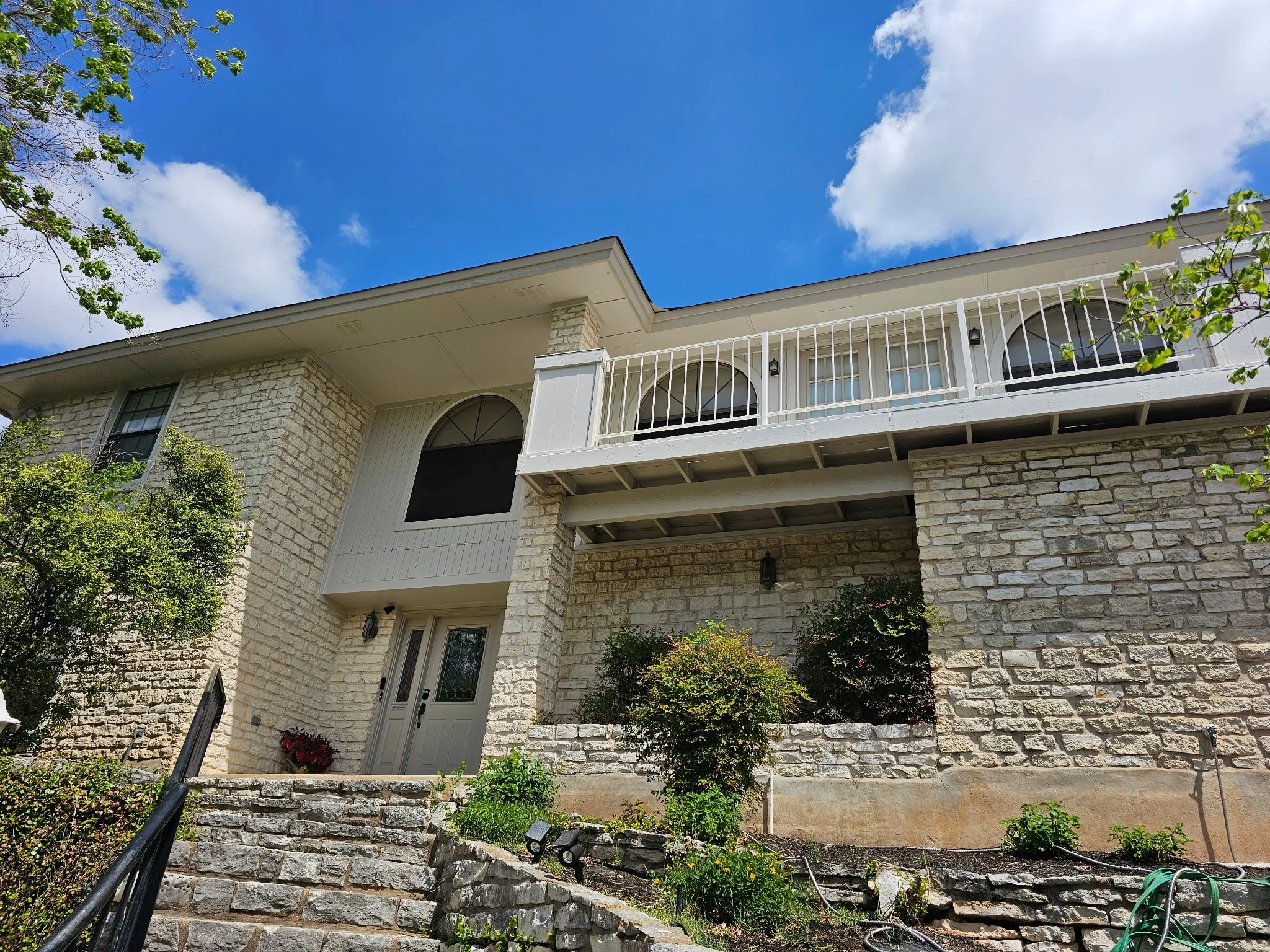 A two-story house with a stone facade, white railing on the balcony, and a front door surrounded by plants and shrubs, with a staircase leading up to the entrance and a bright blue sky with clouds overhead.