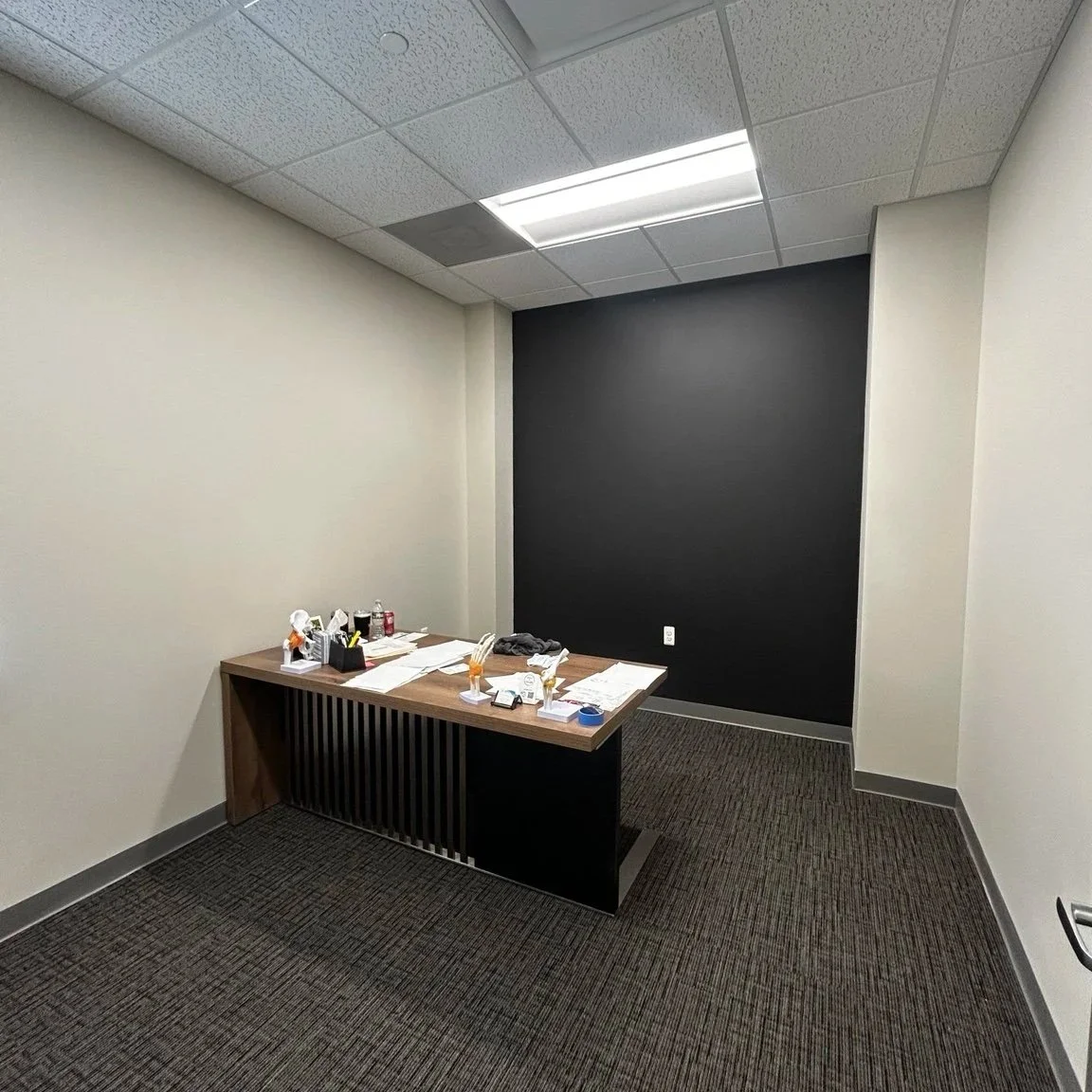 Empty office with a wooden desk and paperwork, items, and chairs, with a black accent wall and fluorescent ceiling lights.
