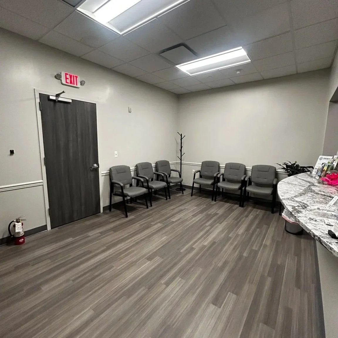 Empty waiting room with a door, six gray chairs, a coat rack, a fire extinguisher, and a curved counter with some magazines.