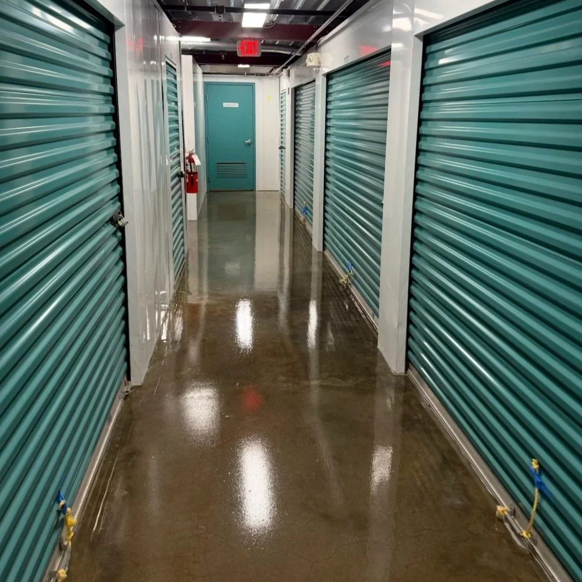 Empty indoor storage unit hallway with green roll-up doors on both sides and a reflective wet floor.