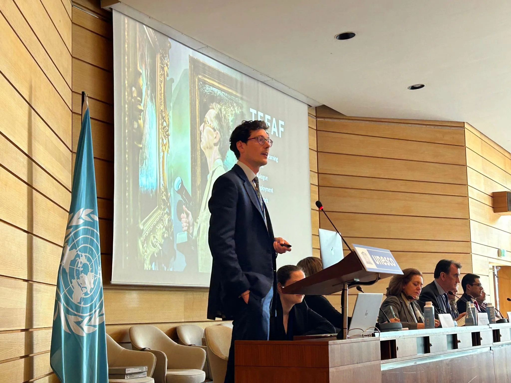 Will Korner speaking at a lectern beside a UN flag at a UNESCO meeting, with a panel seated alongside