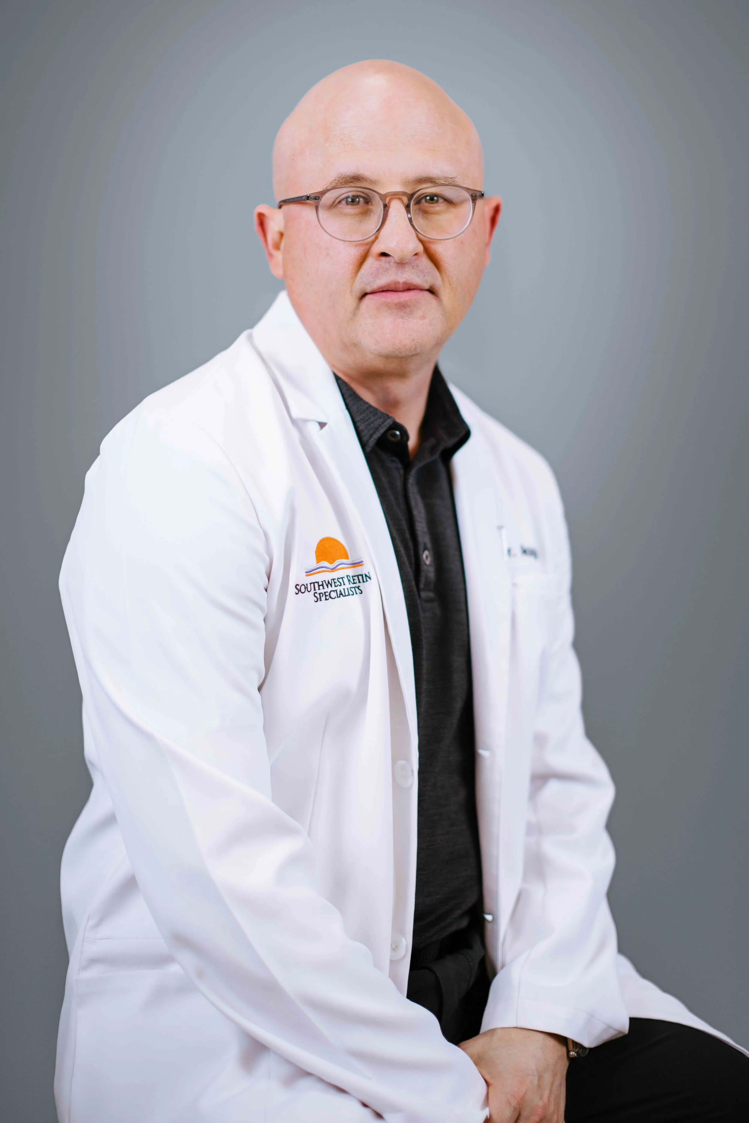 A man wearing glasses and a white doctor's coat with a logo that reads "Southwest Retina Specialists." He is sitting against a gray background, looking directly at the camera with a neutral expression.