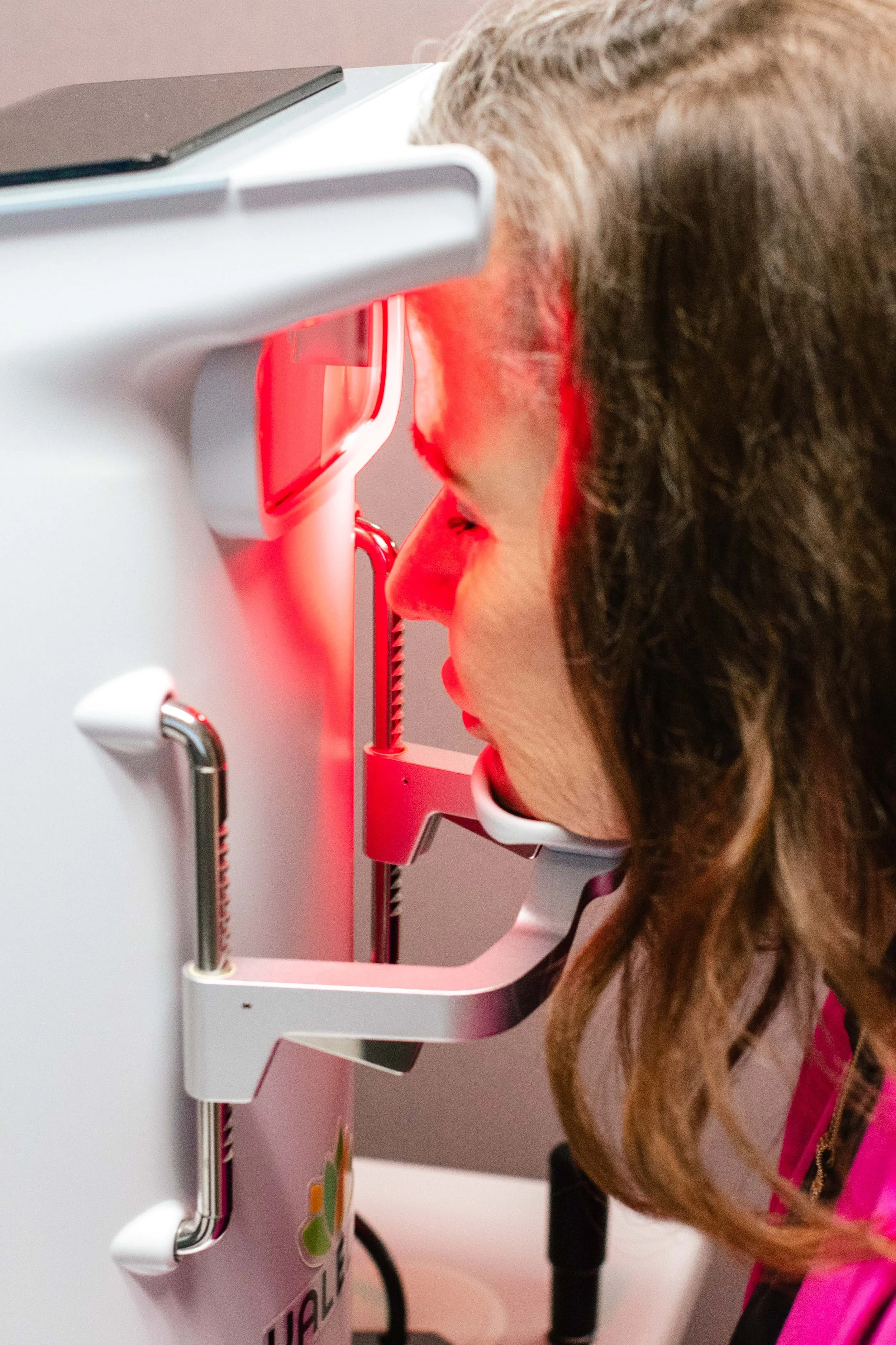 A woman with long brown hair is looking into a medical imaging device that emits red light.