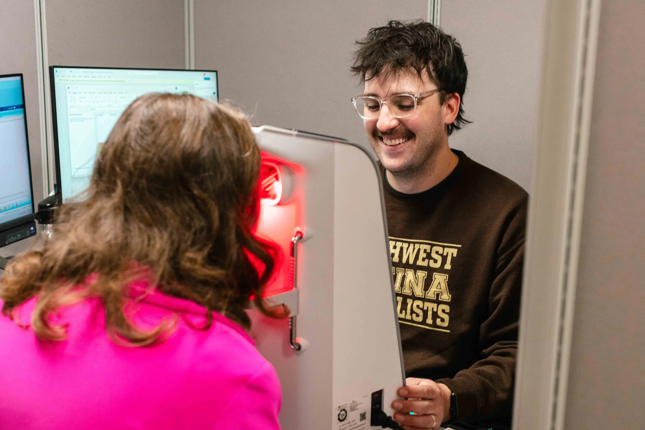 A young man with glasses smiling as he interacts with a patient in a pink jacket during a medical test involving a device emitting red light.