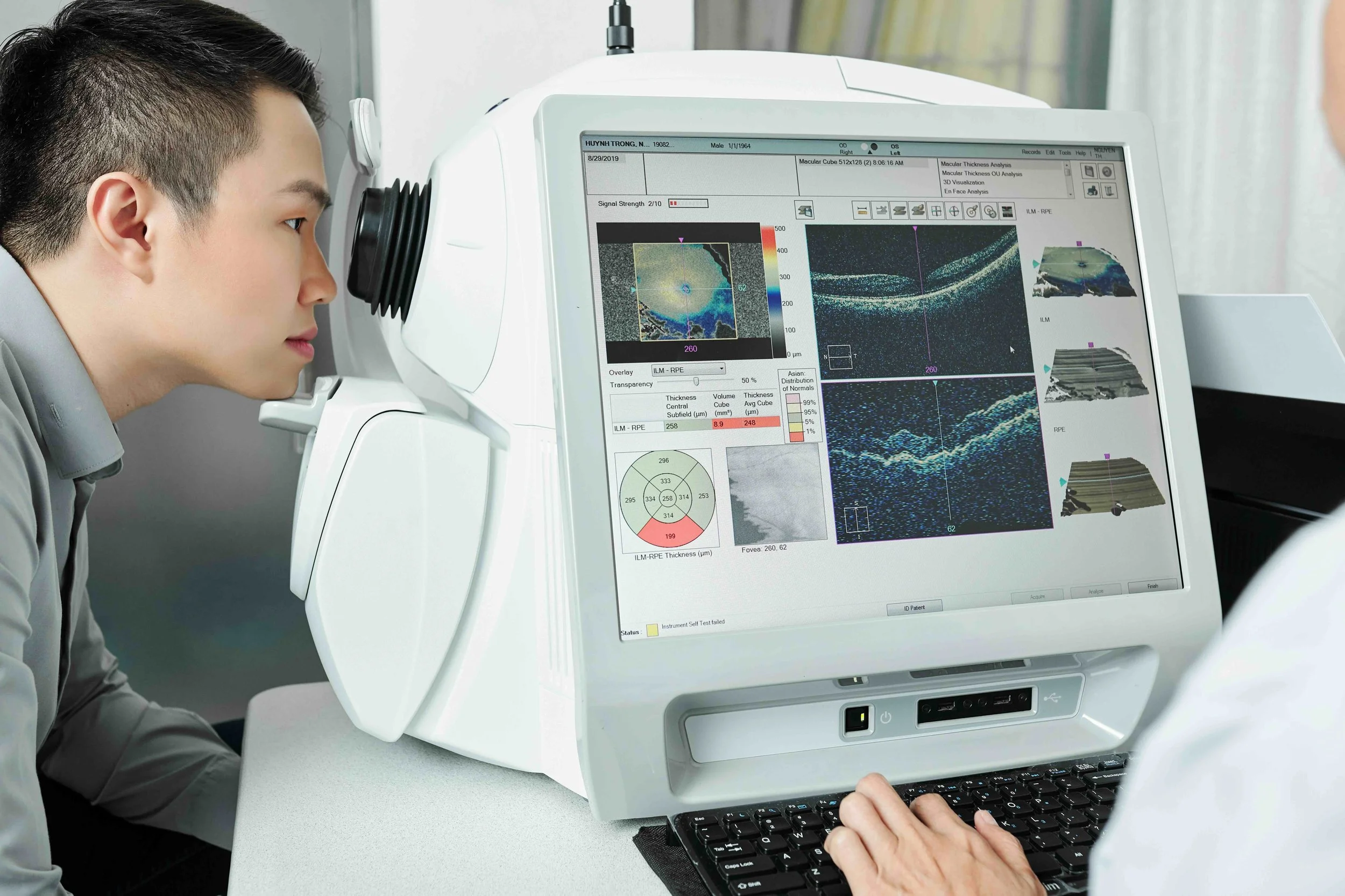 A man with short dark hair looking at a medical machine with a computer screen displaying eye imaging data.
