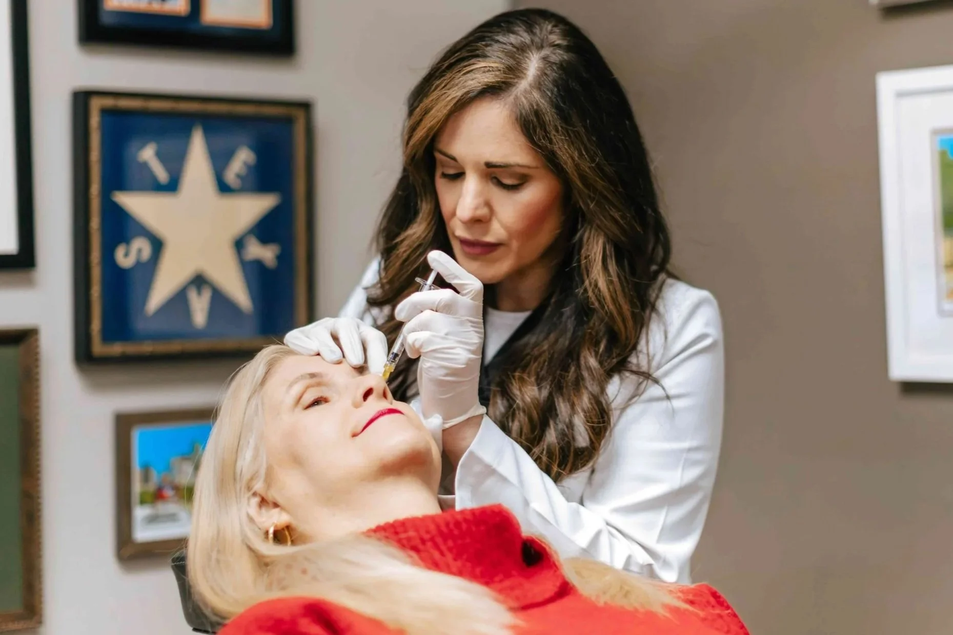 A woman is giving an injection to a woman lying in a chair, with framed art on the wall behind them.